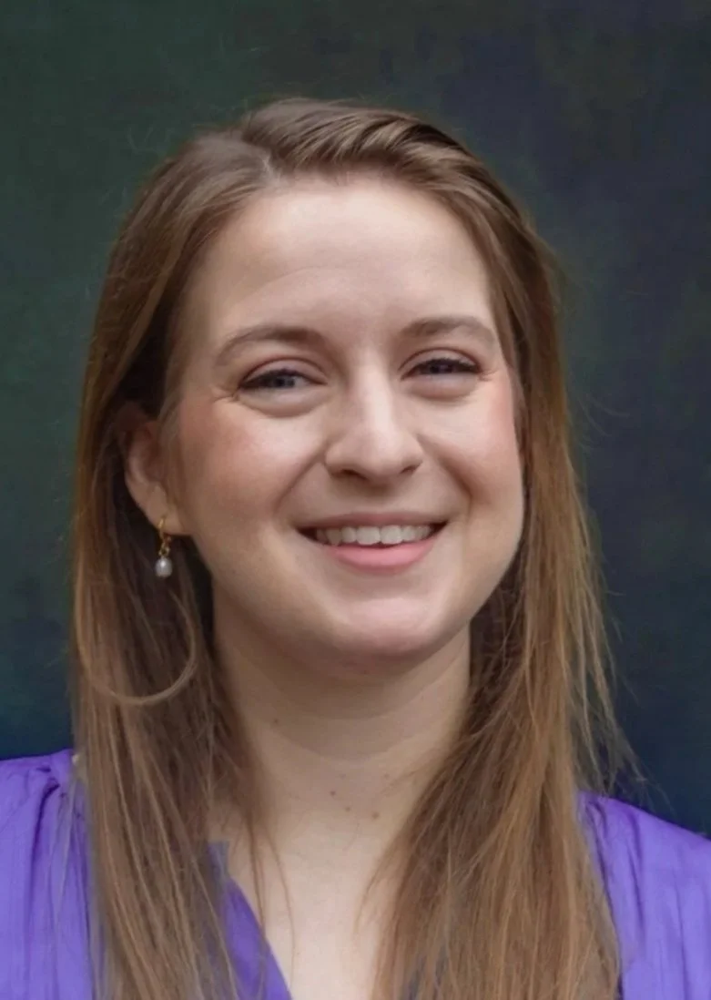 A woman with long light brown hair, smiling, wearing purple clothing and a pearl earring, against a dark, blurry background.