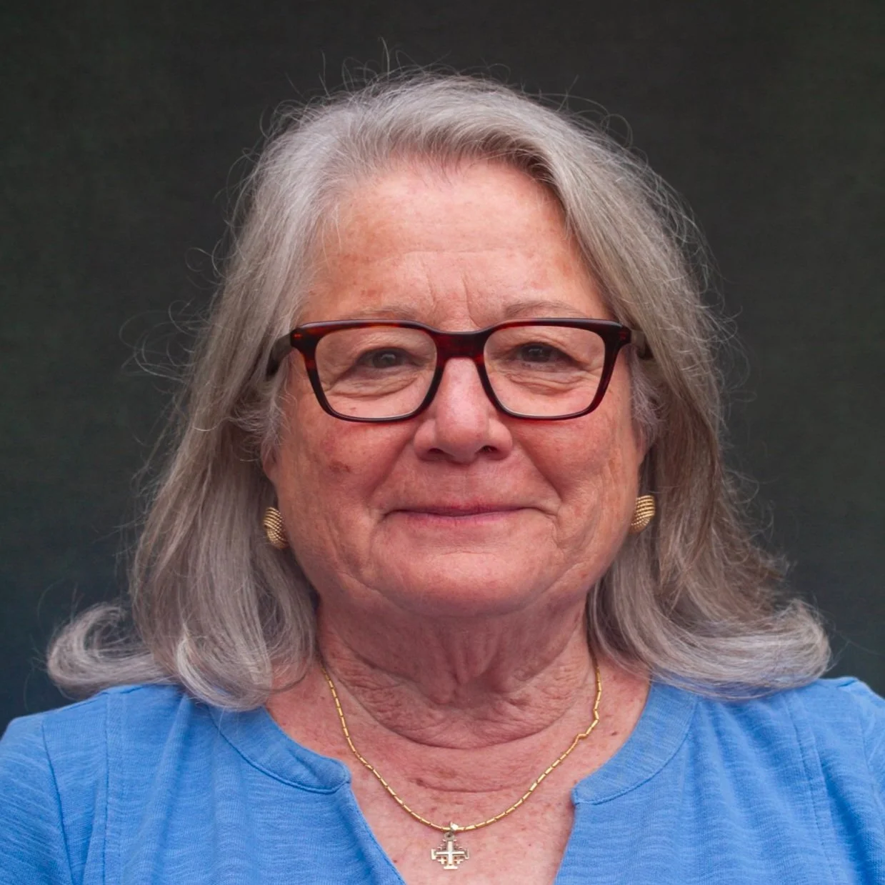 Close-up portrait of a woman with gray hair, wearing glasses and a blue shirt, against a dark background.