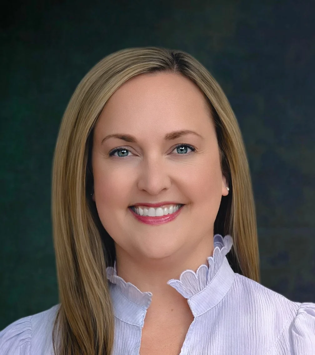 A woman with long blonde hair smiling, wearing a light-colored blouse with a ruffled collar, against a dark background.