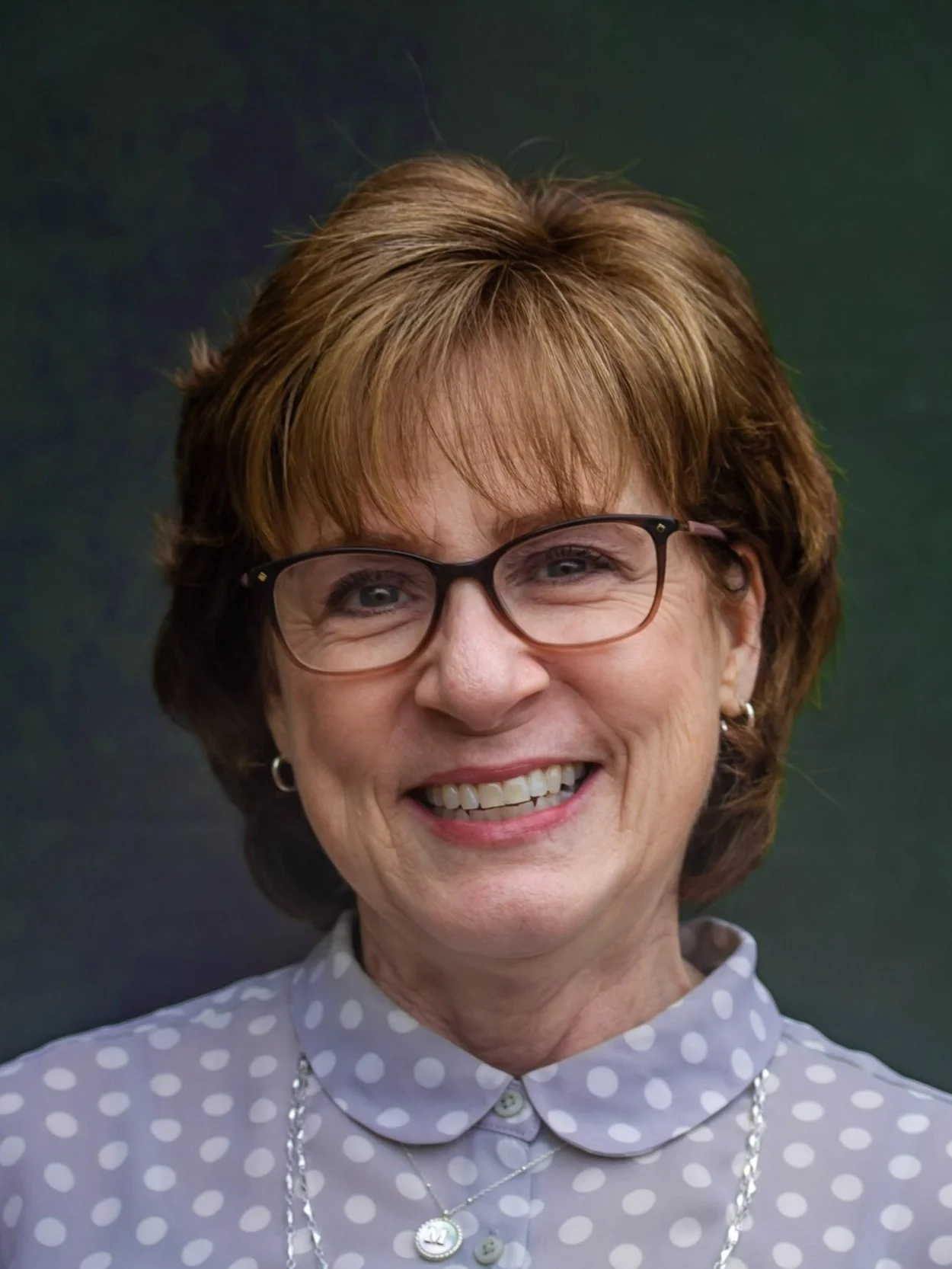 Close-up of a smiling woman with short, reddish-brown hair, wearing glasses, a polka dot shirt, and silver jewelry.