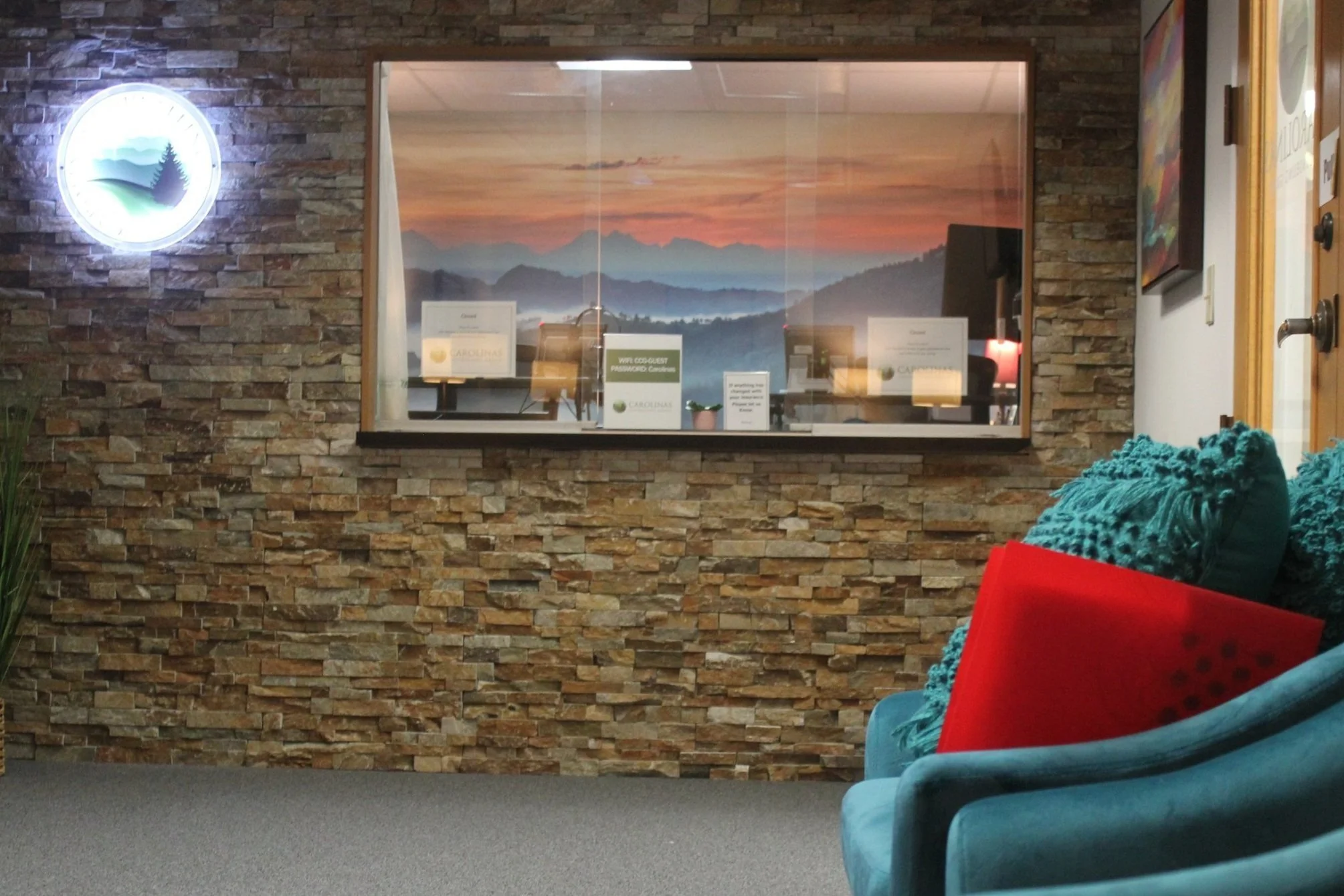Interior of a waiting area with a stone wall, teal armchair with red pillow, and a window showing a sunset over mountains.