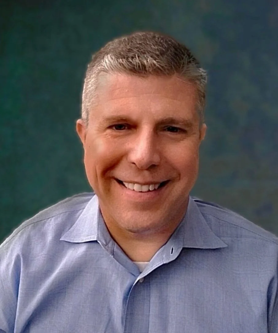 A man with short blond hair and a friendly smile, wearing a light blue dress shirt, outside with leafy shrubbery and trees in the background.