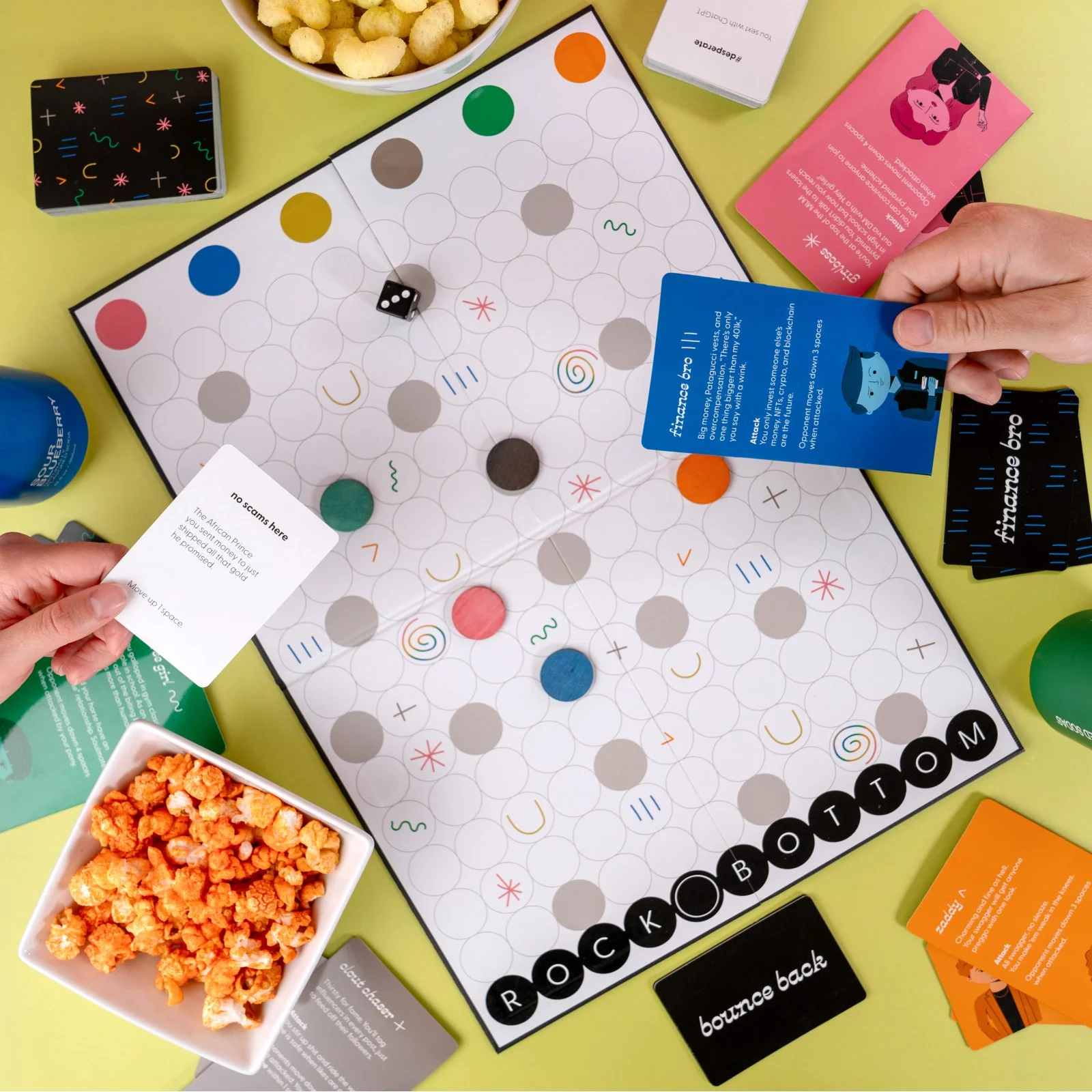 Friends playing Rock Bottom. The image shows a top down view of the game board and pieces surrounded by snacks.