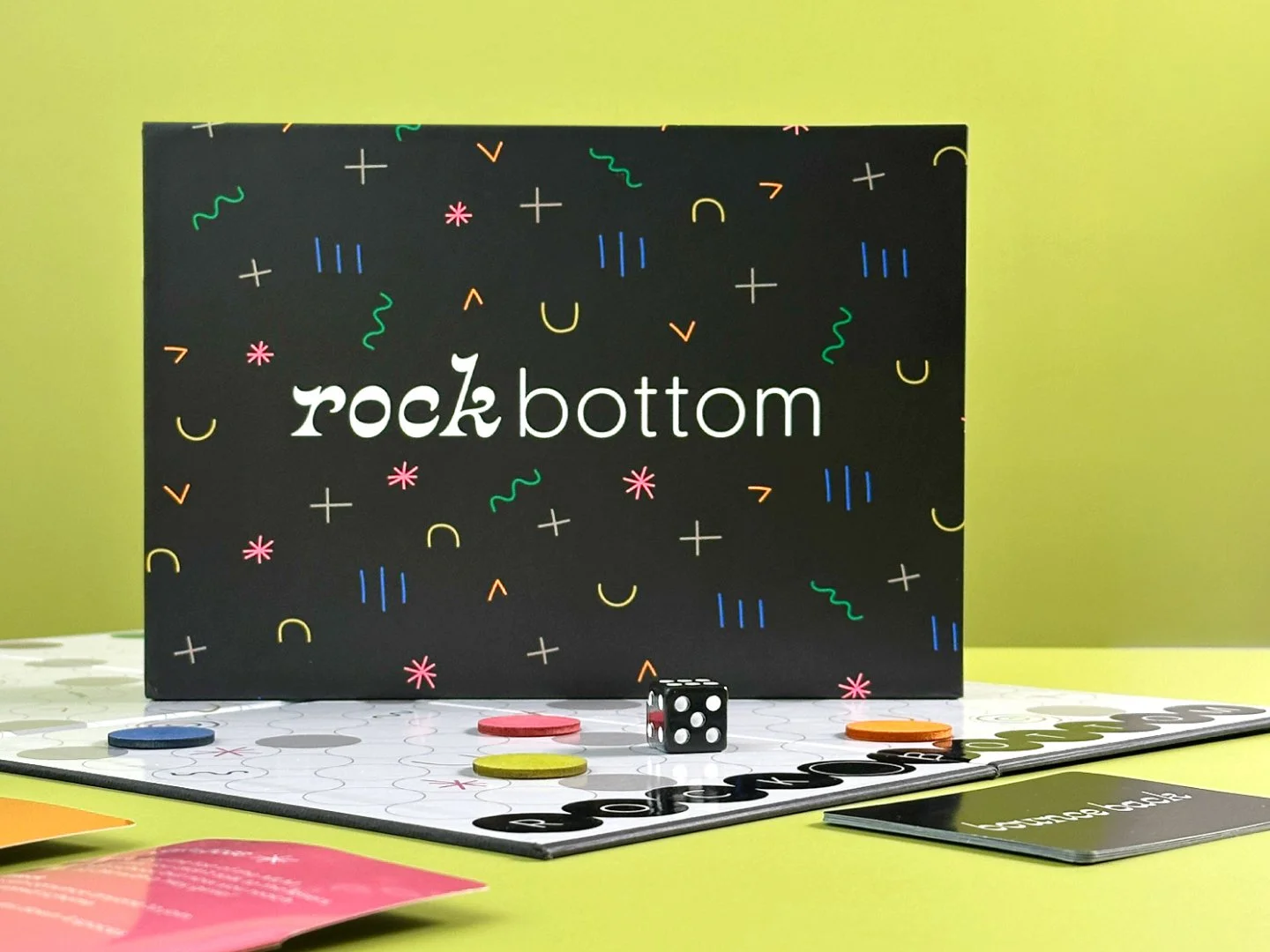 Rock Bottom box sitting on top of the game board with tokens, a die, character cards, and bounce back cards surrounding it.