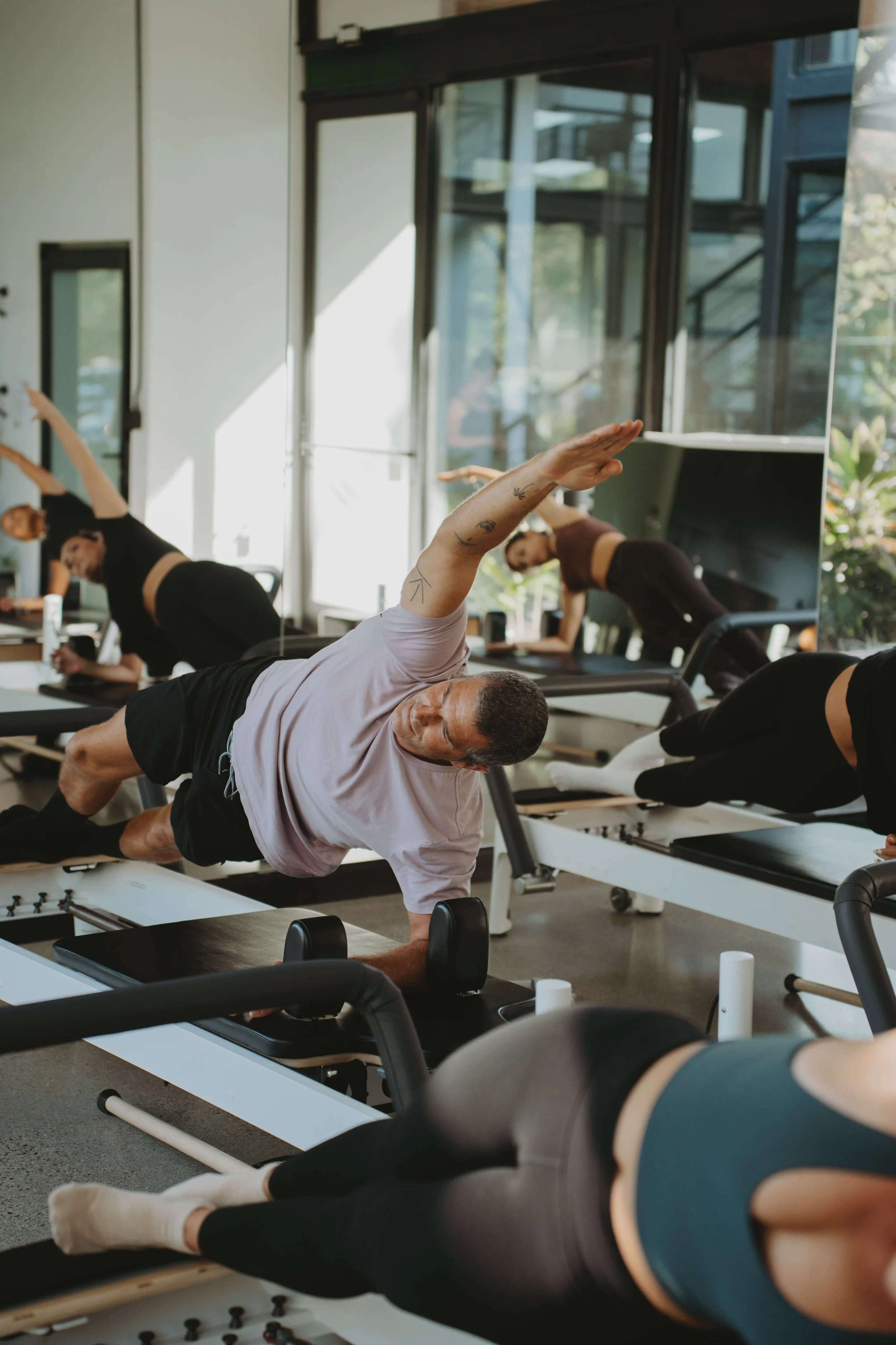People participating in a Pilates class using reformer machines, performing side plank exercises, in a well-lit fitness studio with large windows.