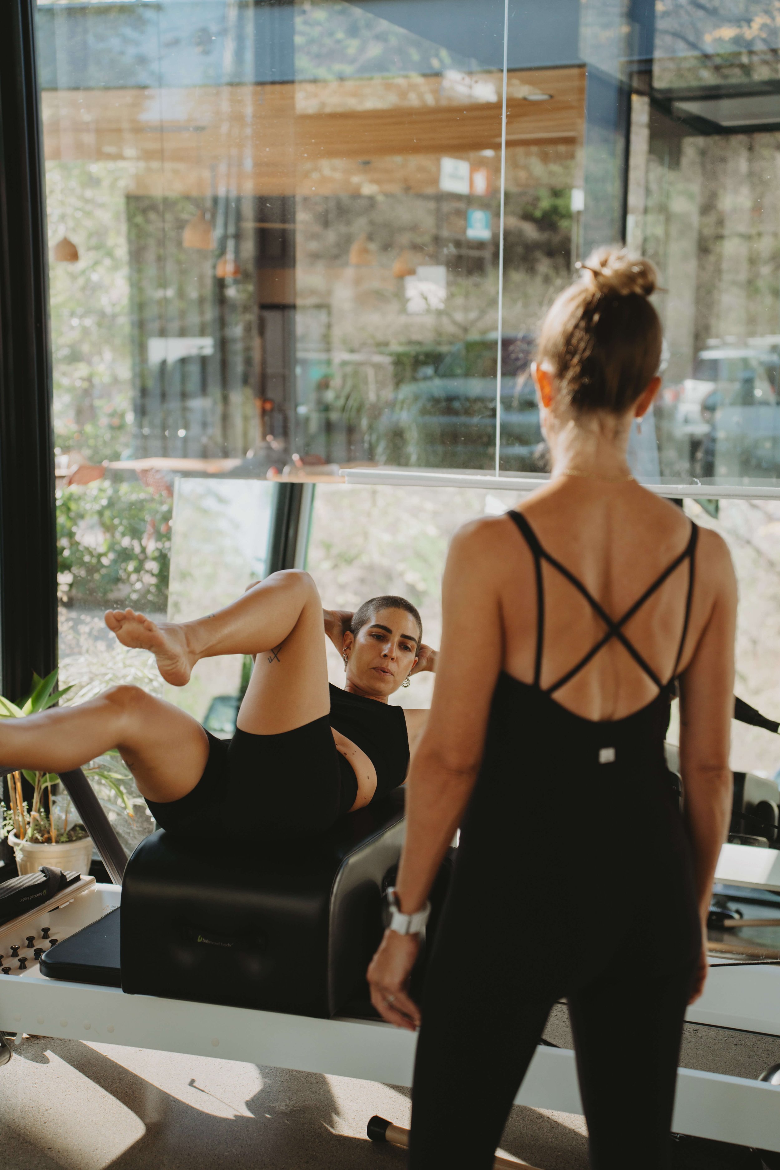 Two women in a gym, one lying on exercise equipment doing leg raises, the other standing with her back to the camera, both in workout clothes.