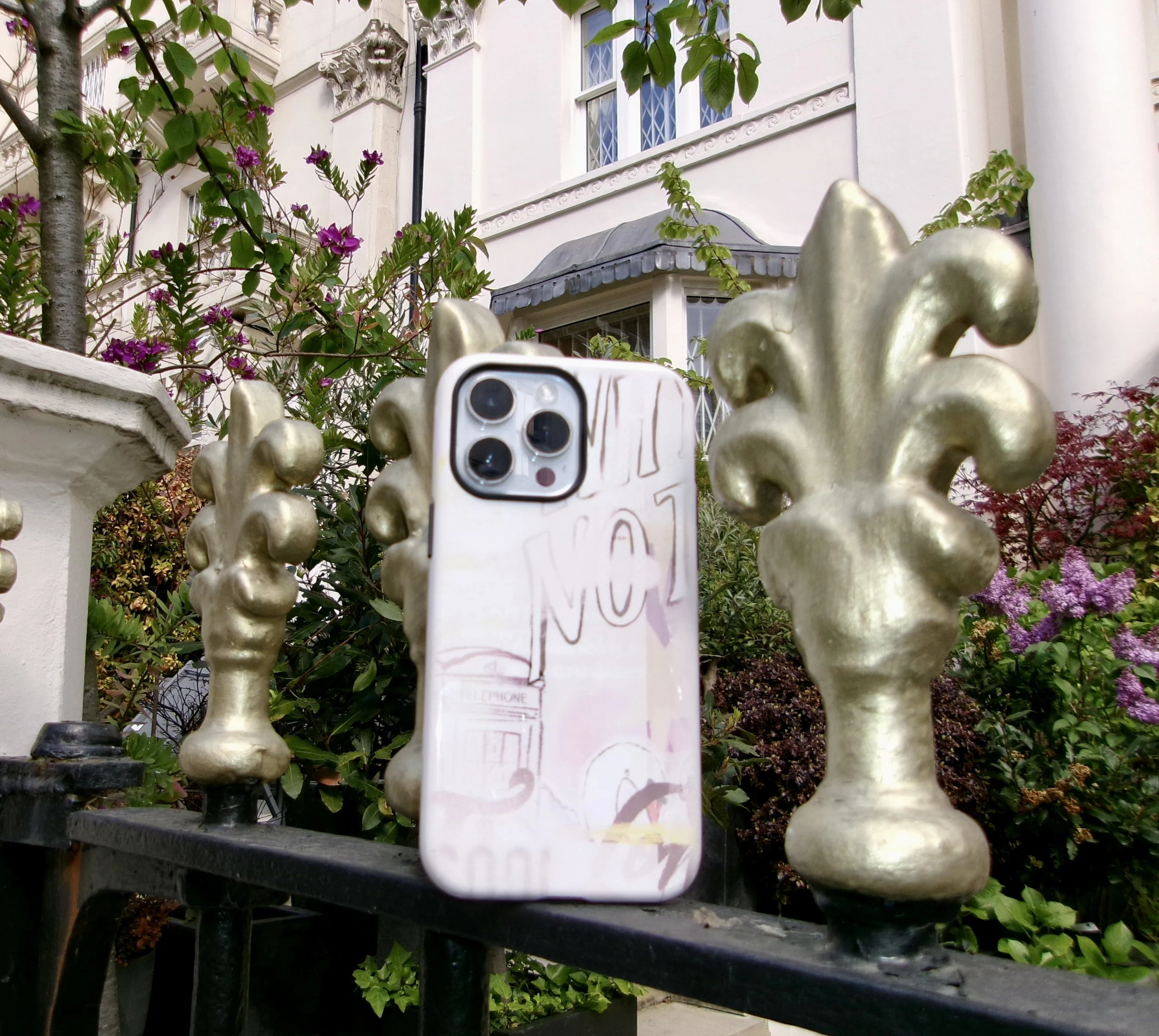 A golden decorative fence with a white smartphone attached, taking a panoramic photo of a white house with ornate window frames, surrounded by blooming purple flowers and green foliage.