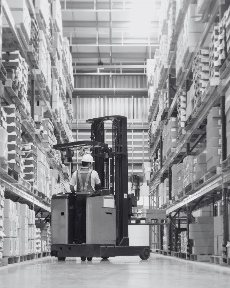 Warehouse career transition support. A worker operating a forklift in a warehouse aisle surrounded by tall shelves filled with boxes.