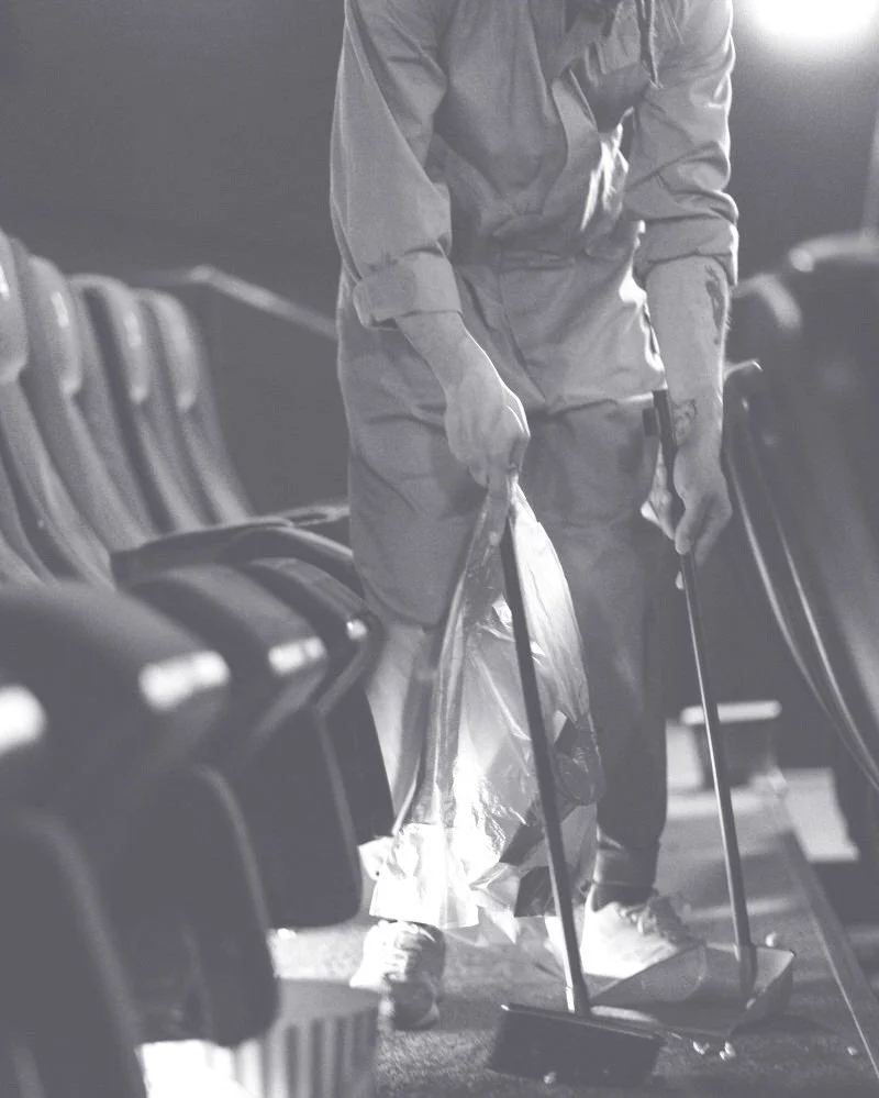 A person wearing a jumpsuit and sneakers cleaning or setting up chairs with a tool in a dimly lit indoor space.