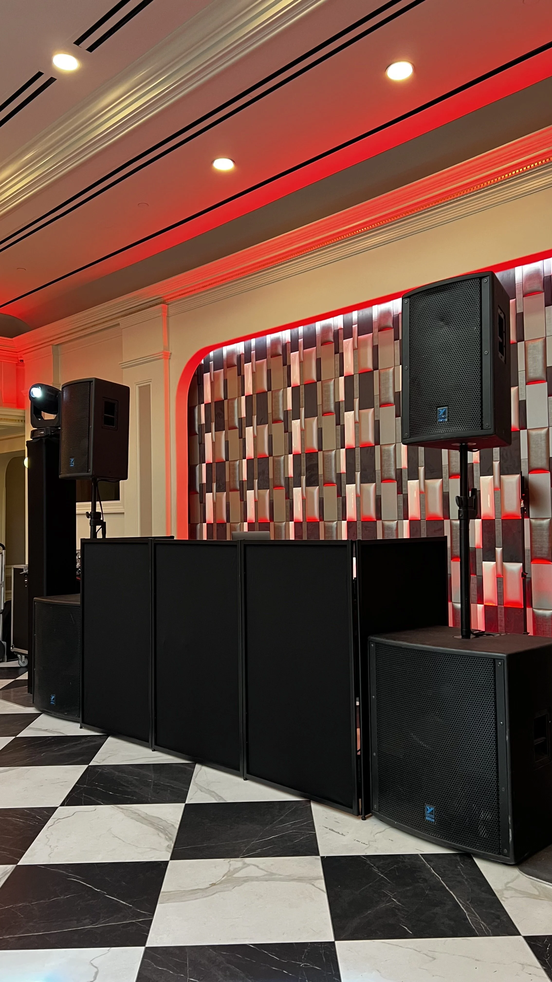 Sound system with multiple large black speakers set up on a stage in an indoor venue with a decorative wall and checkered black and white floor.