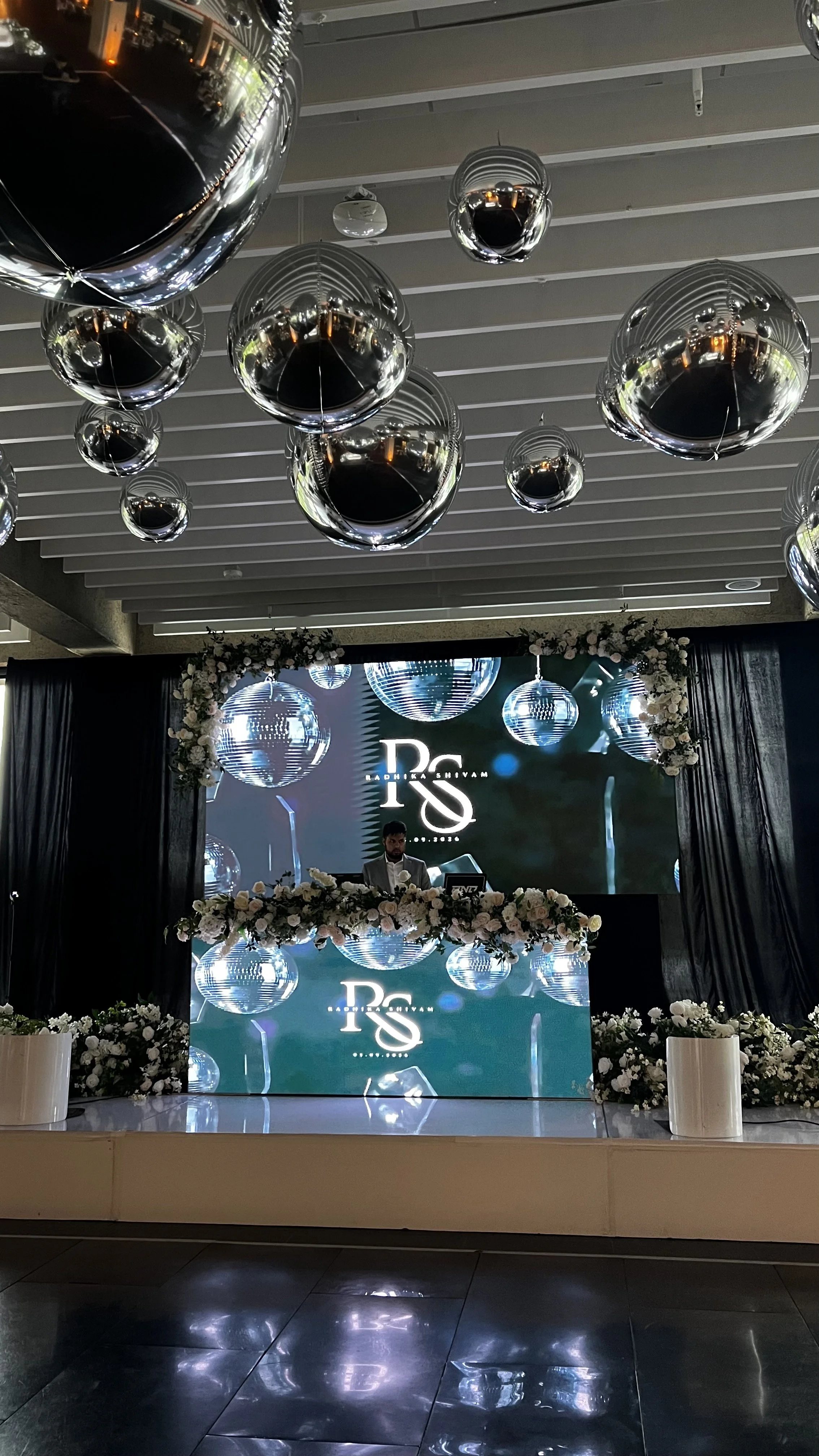 A decorated stage with hanging reflective balloons, floral arrangements, and a large digital screen displaying the initials 'D' and 'S' for a celebration event.