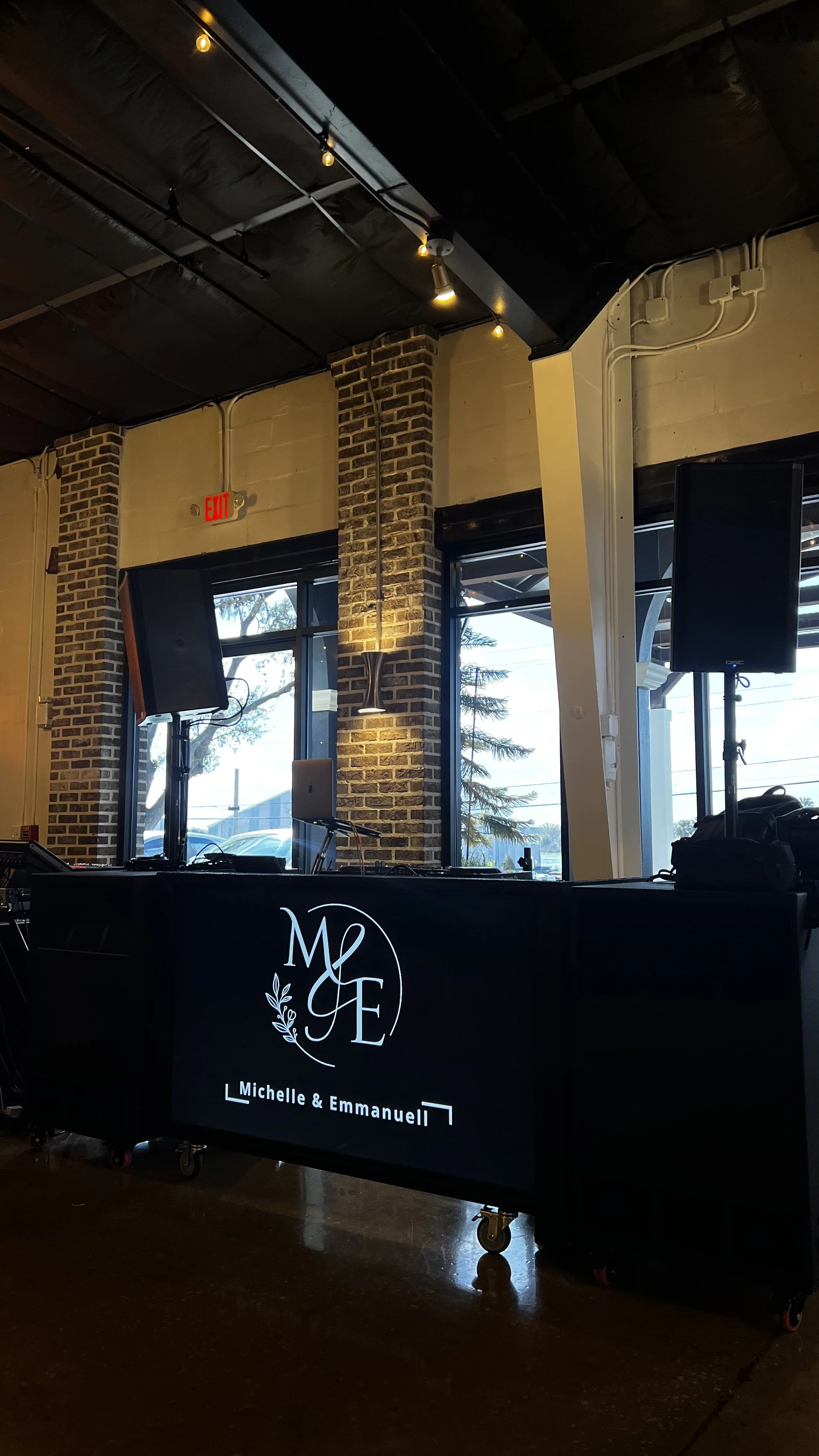 A DJ setup with two speakers, a laptop, and a DJ table with a black cloth displaying white text and a decorative logo, in an indoor venue with large windows, exposed brick pillars, and ceiling lights.