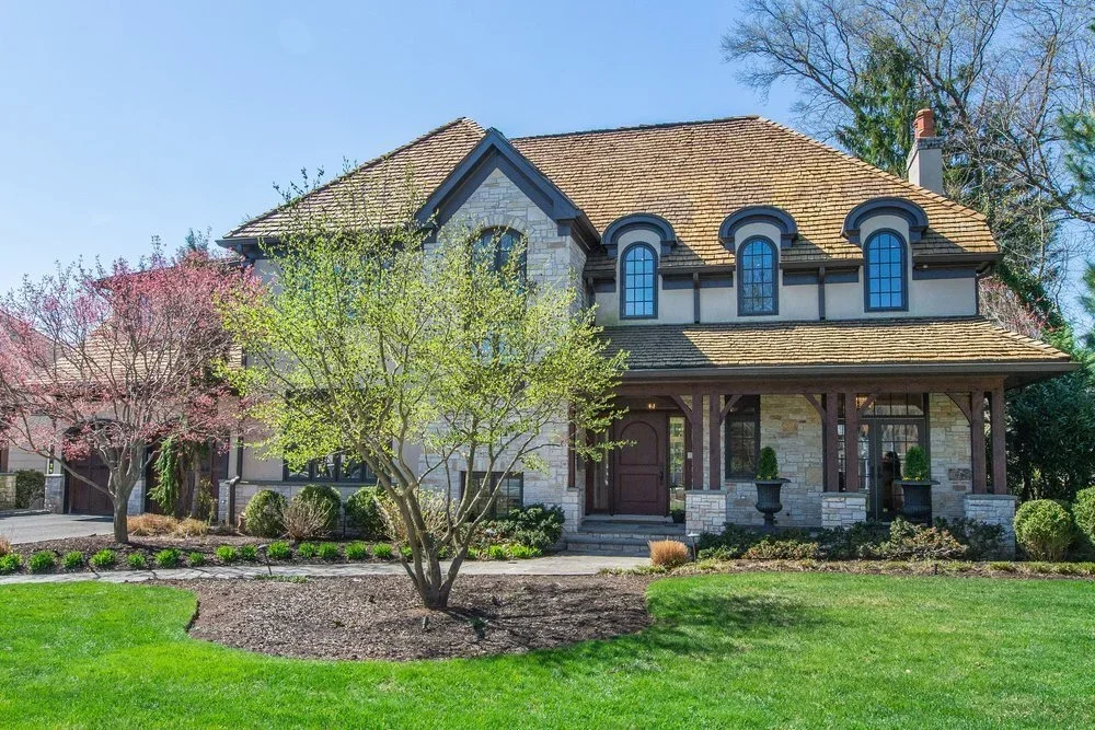 A large two-story house with a mix of stone and wood exterior, arched windows, a covered porch, and a well-maintained front yard with grass, trees, and shrubs.