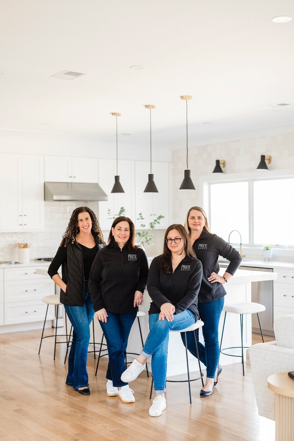 Four women in casual outfits pose and smile together in a bright, modern kitchen with white cabinets and pendant lights.