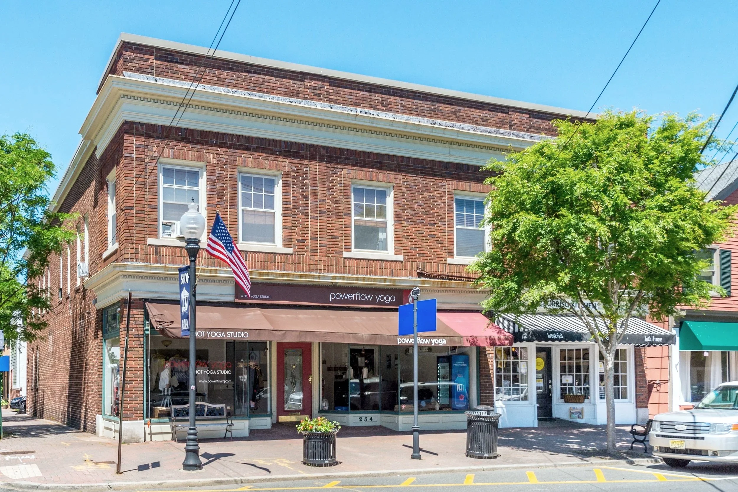 Two-story brick building with storefronts, one named 'powerflow yoga', storefronts have awnings, trees on sidewalk, American flag on lamppost, parked car, blue sky.