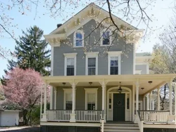 A large gray house with a front porch, multiple windows, and a pitched roof, surrounded by trees, including a pink flowering tree.