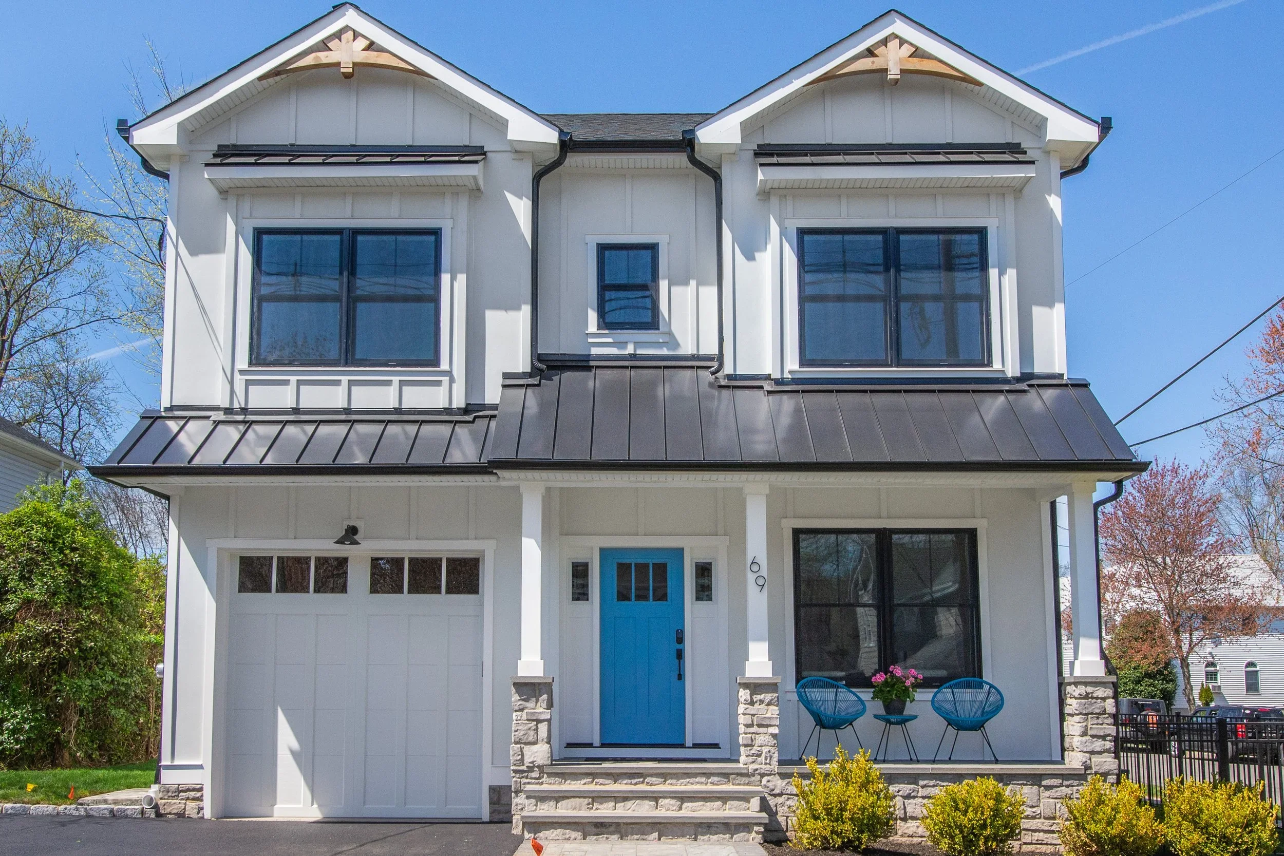 Front view of a white two-story house with a black metal roof, black window frames, a bright blue front door, and a small front porch with two blue chairs and a flowerpot. The house has stone accents at the base, and the sky is clear and blue.
