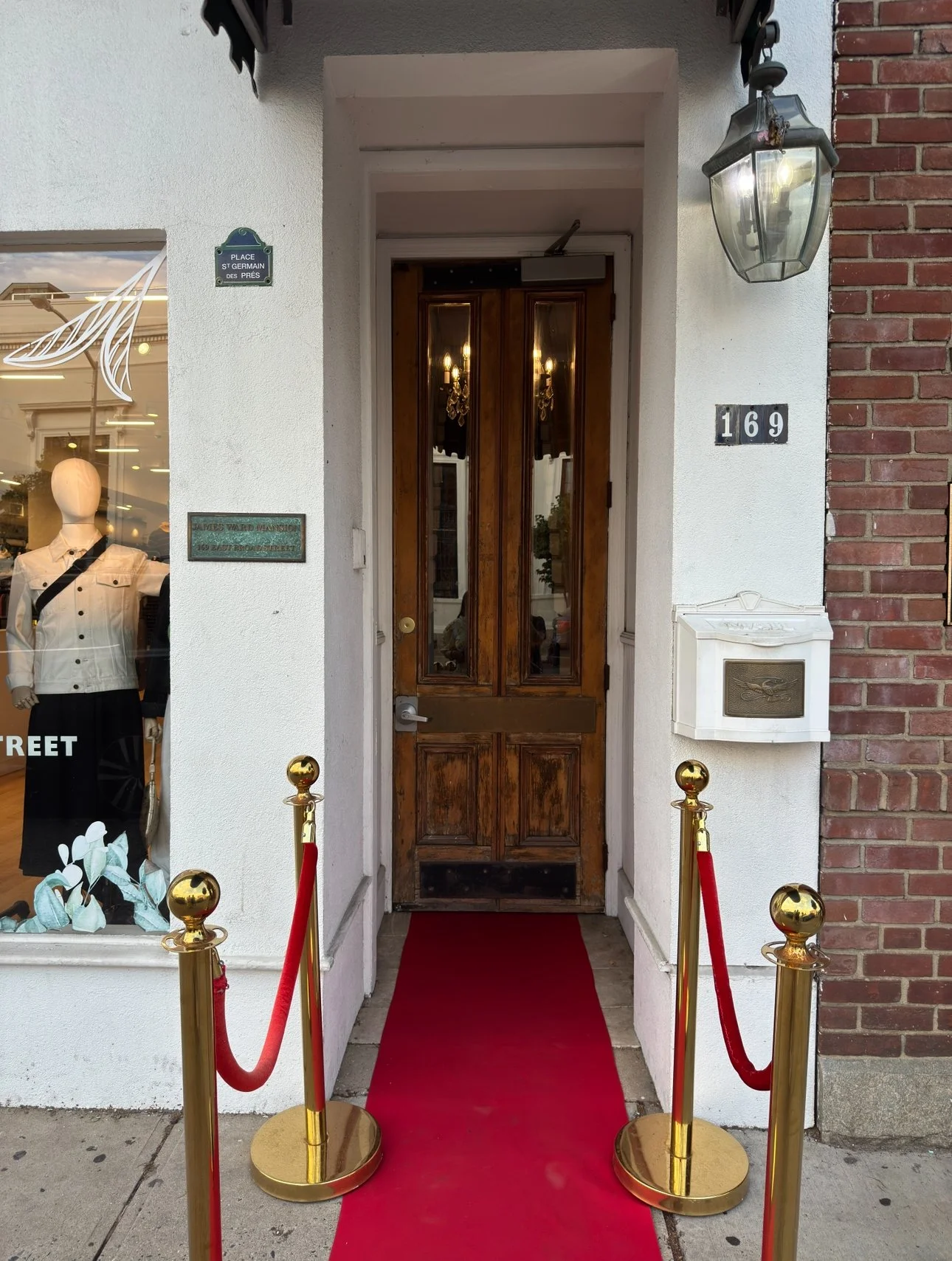 A doorway with a red carpet, gold stanchions with red velvet ropes, and a wooden door. To the left, there is a store window with mannequins displaying clothing. The building has a large glass lantern hanging on the right side of the door, and a mailbox on the wall to the right.