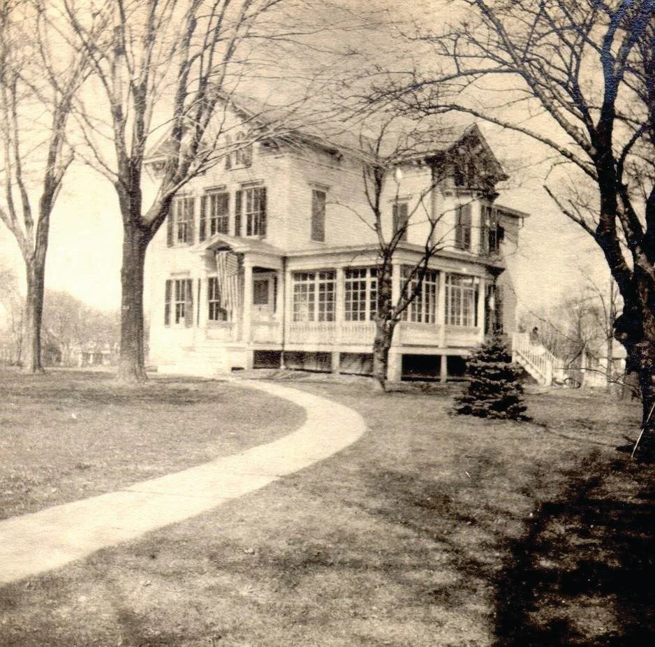 An old black and white photo of a large, old-fashioned house with multiple stories, surrounded by leafless trees and a curved walkway leading to the front porch.