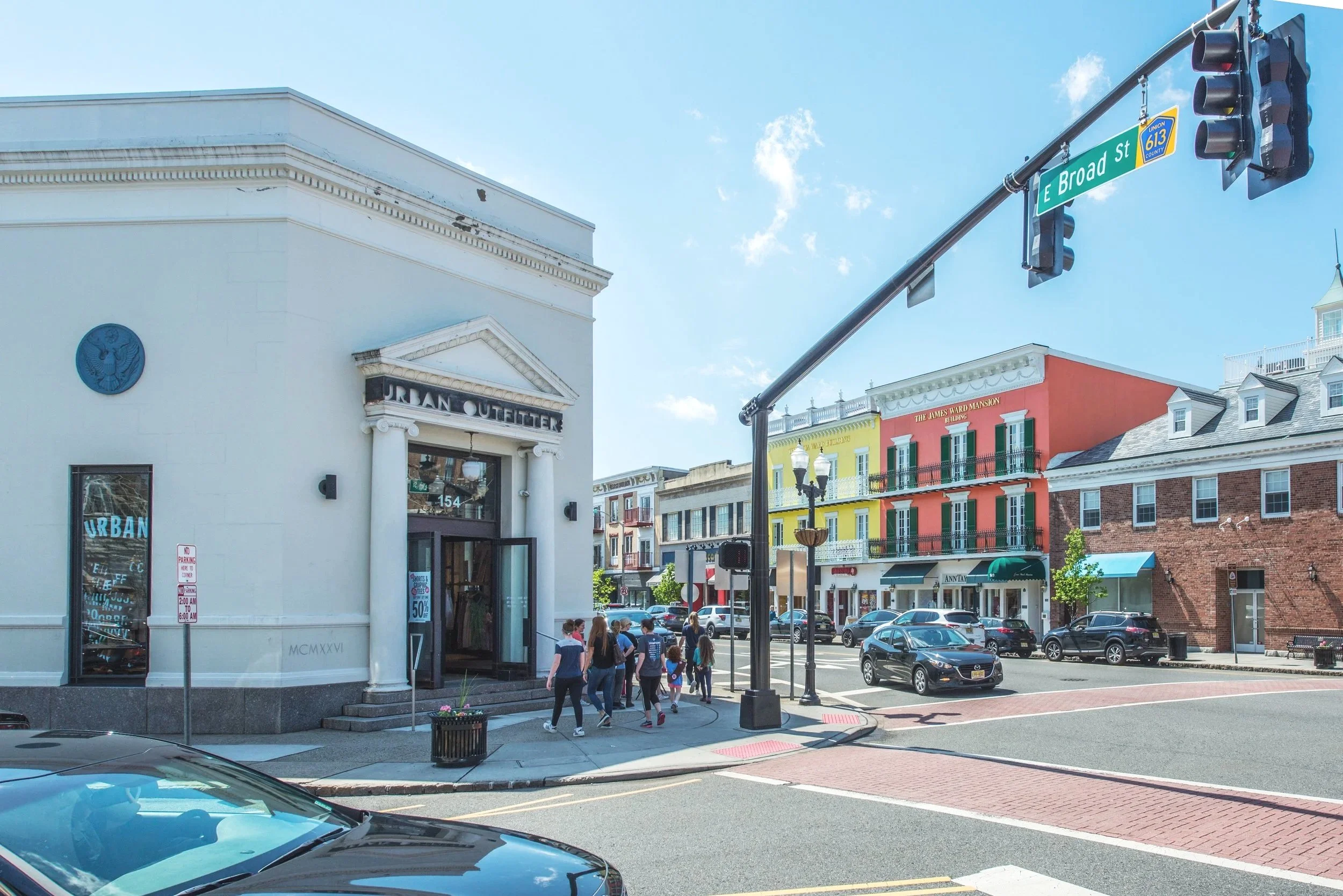 A busy street corner with storefronts, including the Urban Outfitters, and pedestrians walking on the sidewalk under a blue sky with some clouds. Traffic lights and street signs are visible, with the street named E Broad Street.