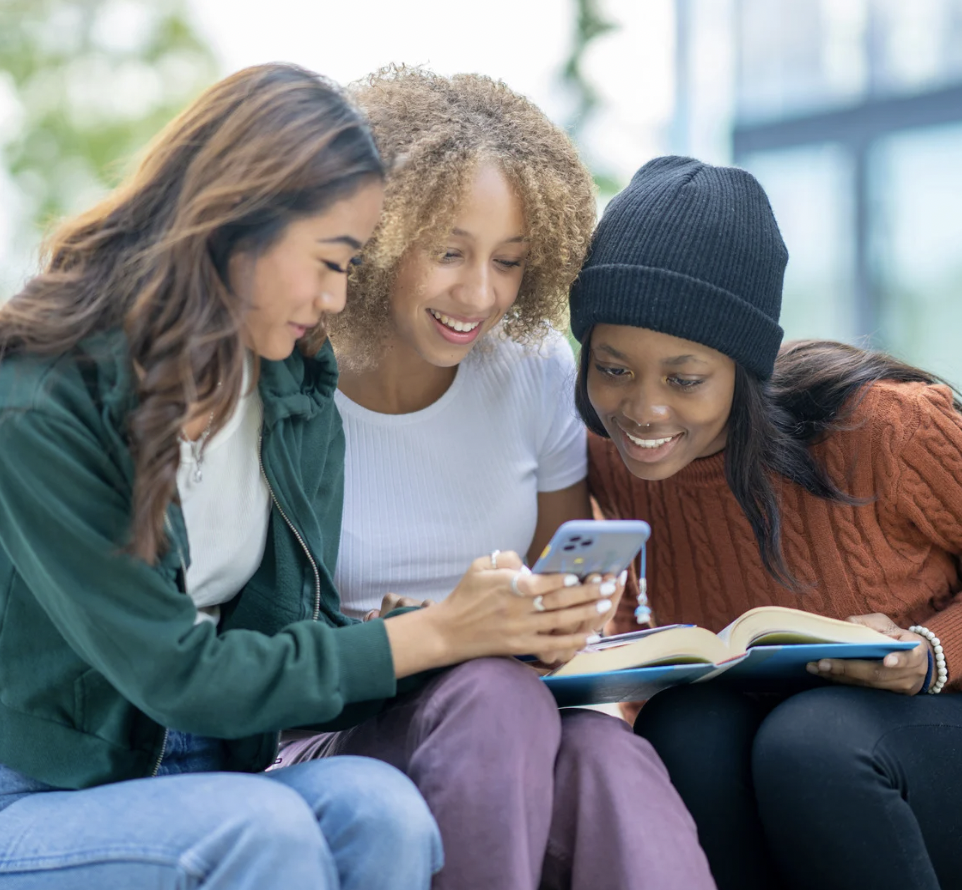 Three young women sitting close together outdoors, looking at a smartphone and smiling, with open books on their laps.