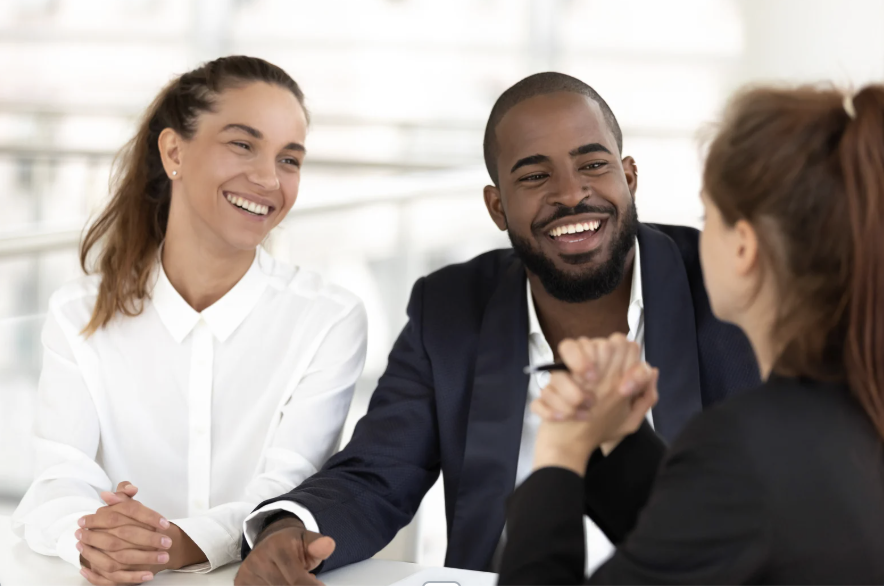 Three diverse businesspeople engaged in a conversation, smiling and holding hands on a white table in a bright office.