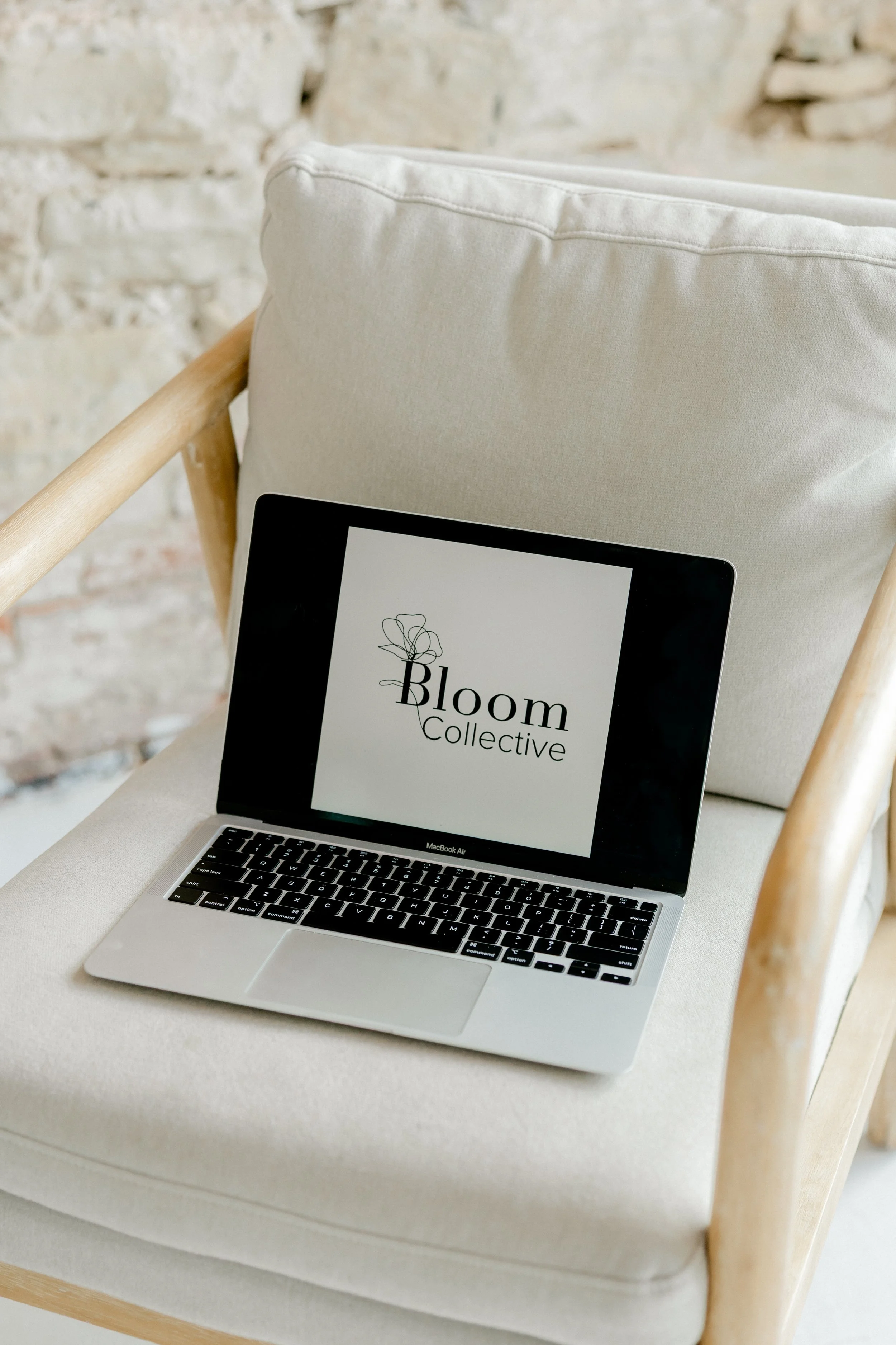 A laptop with a logo for Boom Collective on the screen, placed on a beige upholstered chair with a wooden frame, against a brick wall background.