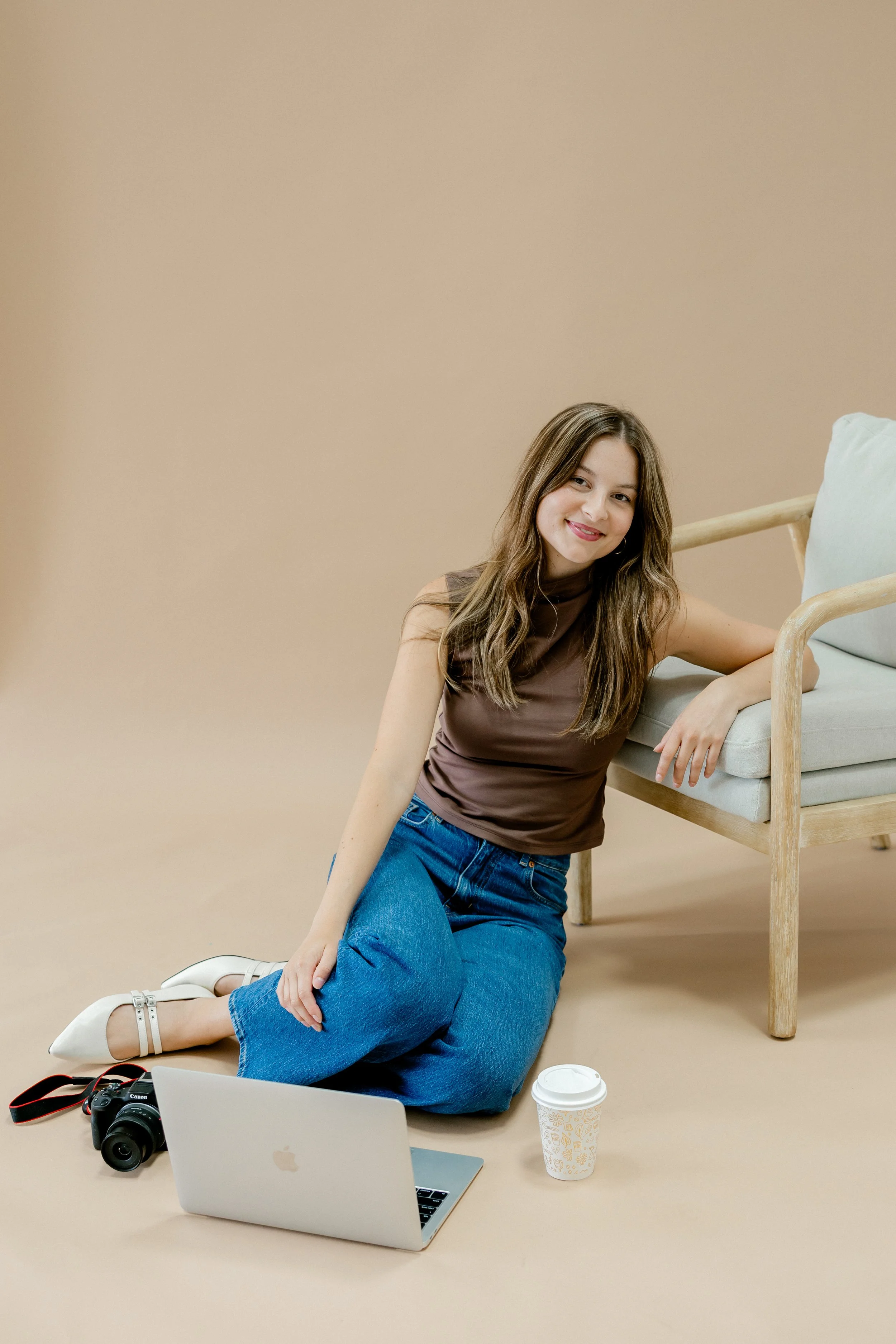 A woman with long brown hair is sitting on the floor, smiling at the camera. She is dressed in a sleeveless brown top and blue jeans. Next to her are a laptop, a camera, and a disposable coffee cup. There is a beige armchair with light pillow in the background.