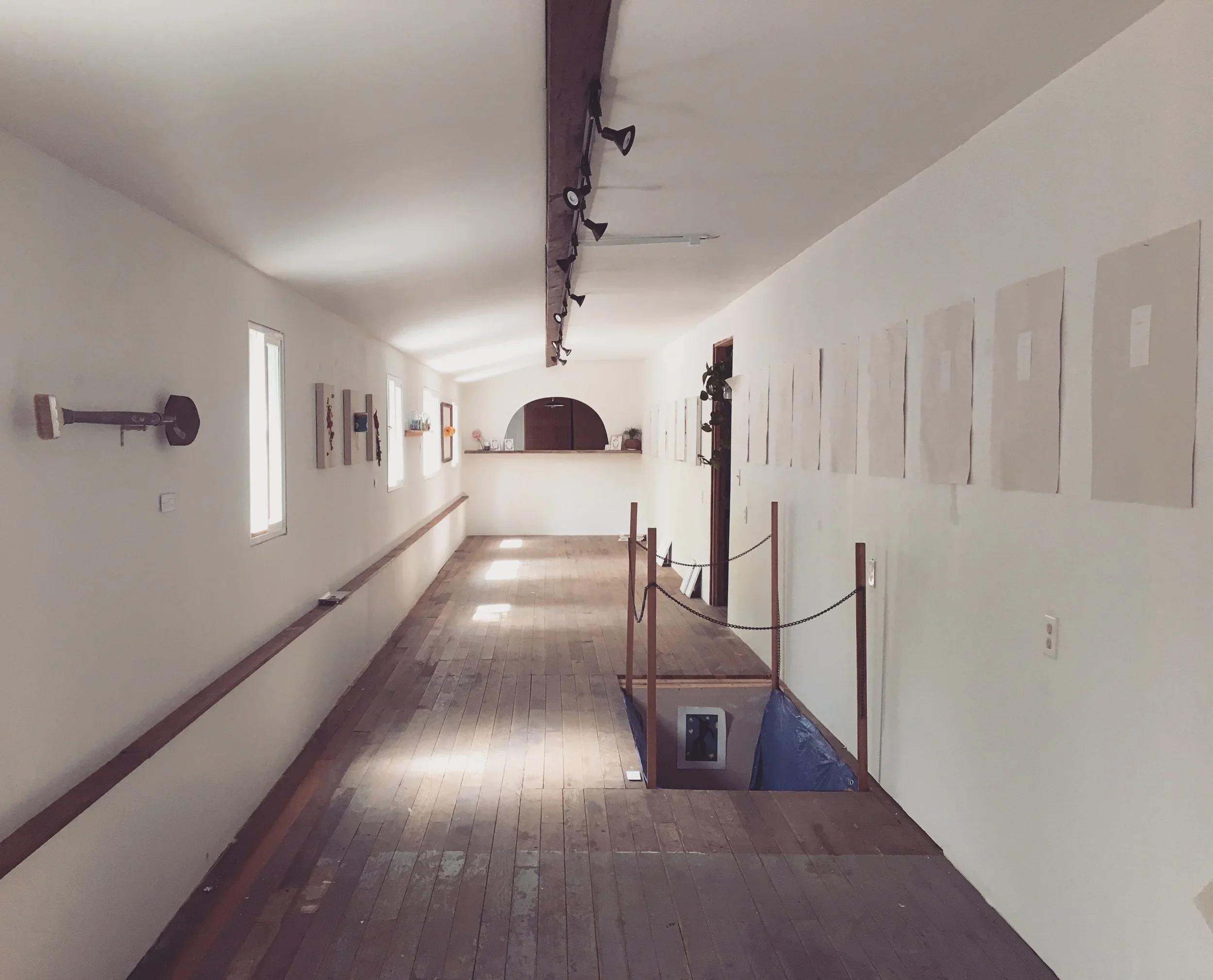 Long corridor in an art gallery with wooden floors, artwork on the walls, and a staircase leading down with a protective railing.