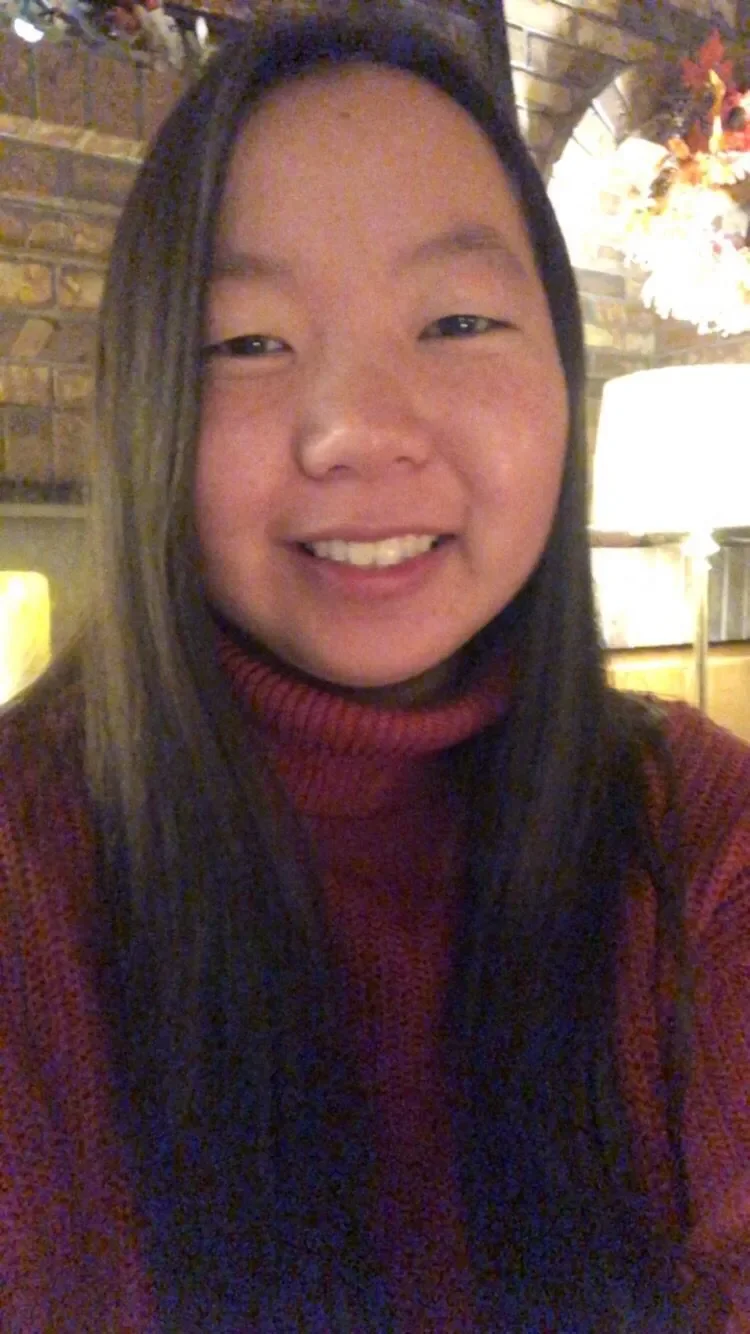 A young woman with long dark hair smiling, wearing a red turtleneck sweater, sitting indoors with a brick wall and a lamp in the background.