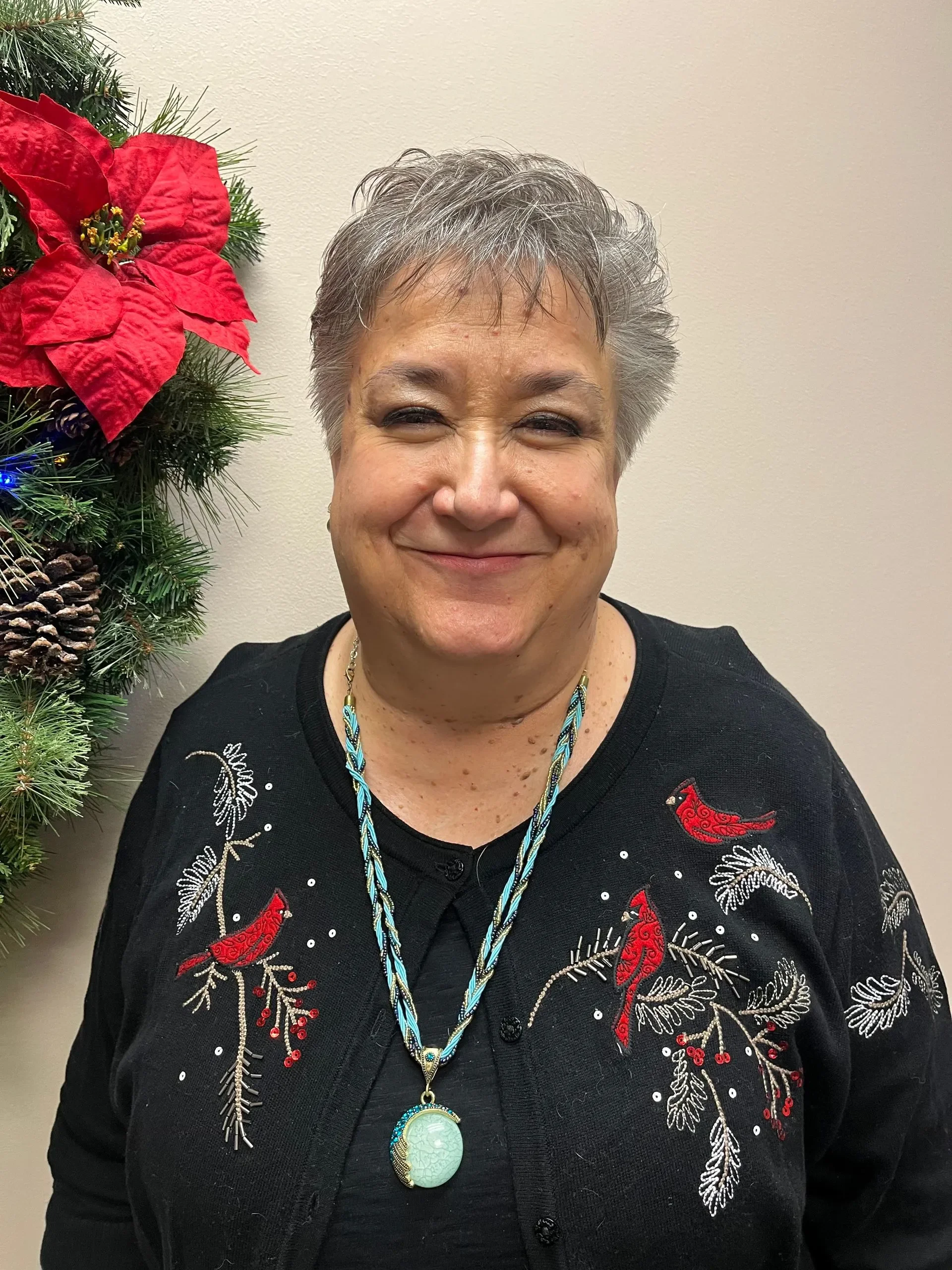 A smiling woman with short gray hair wearing a black Christmas sweater with red bird and holiday decorations, standing next to a festive decor with poinsettia and pine branches.