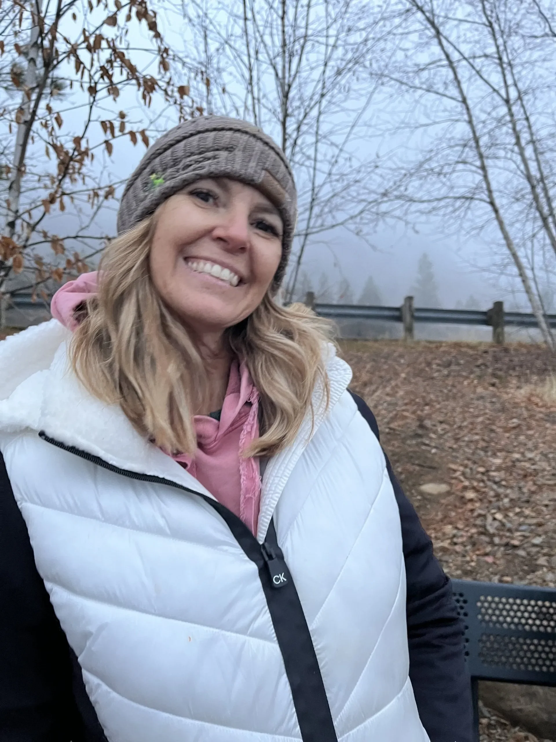 A smiling woman with blonde wavy hair wearing a brown knit beanie, pink hoodie, and white quilted vest outdoors on a foggy day, with leafless trees and a wooden fence in the background.