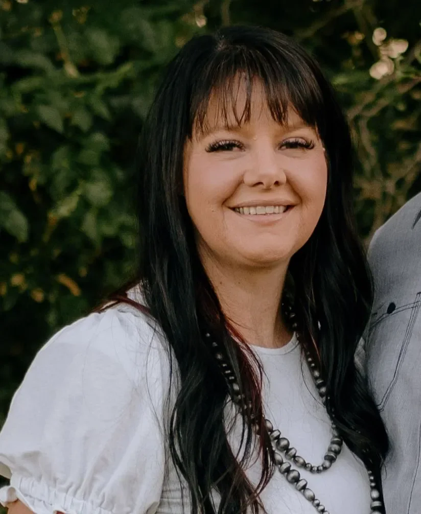 A smiling woman with long black hair and bangs, wearing a white top and a beaded necklace, standing outdoors with greenery in the background.