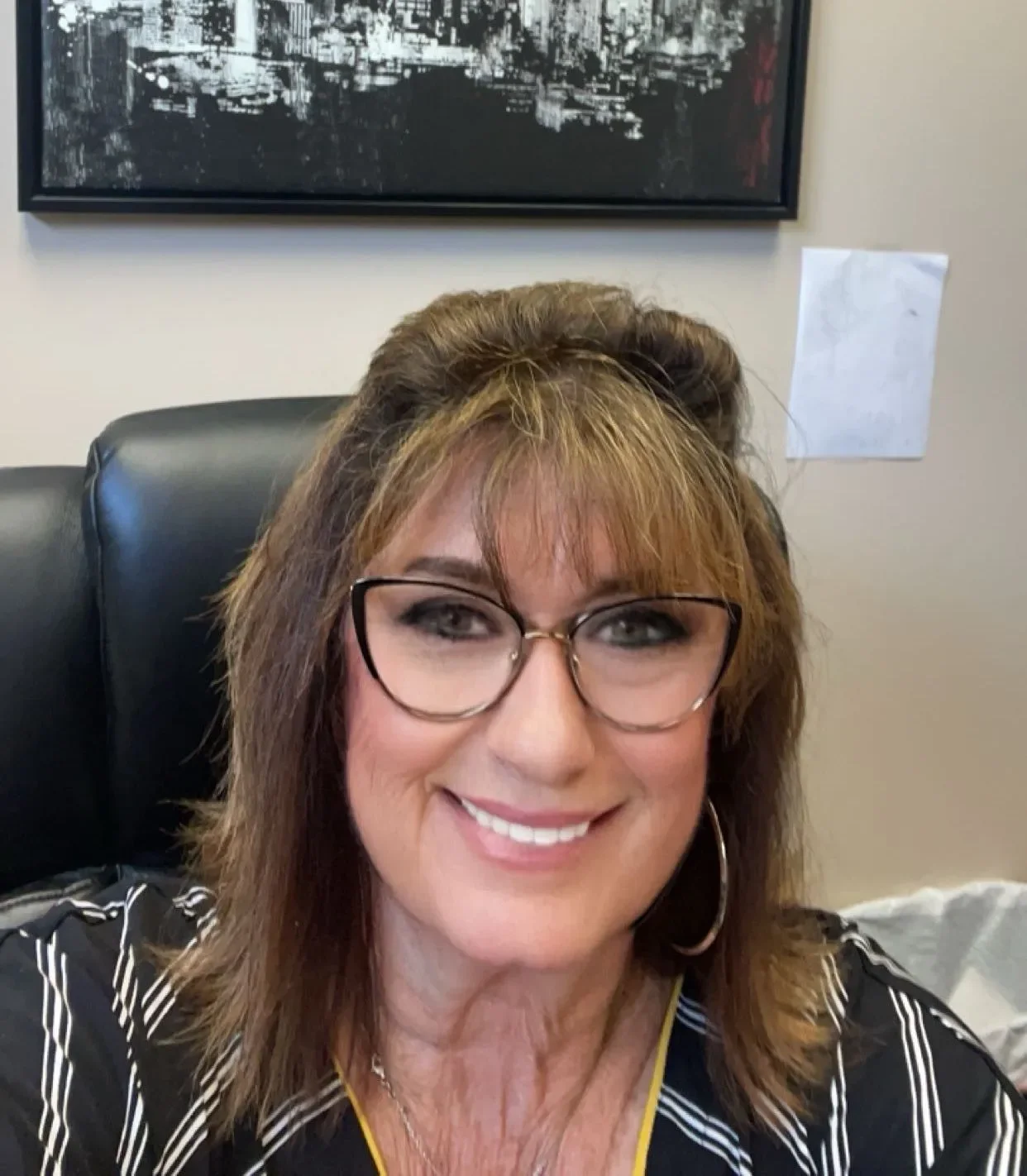 A smiling woman with glasses, wearing a black and white striped top, sitting in an office chair in front of a beige wall with a black and white cityscape picture hanging on it.