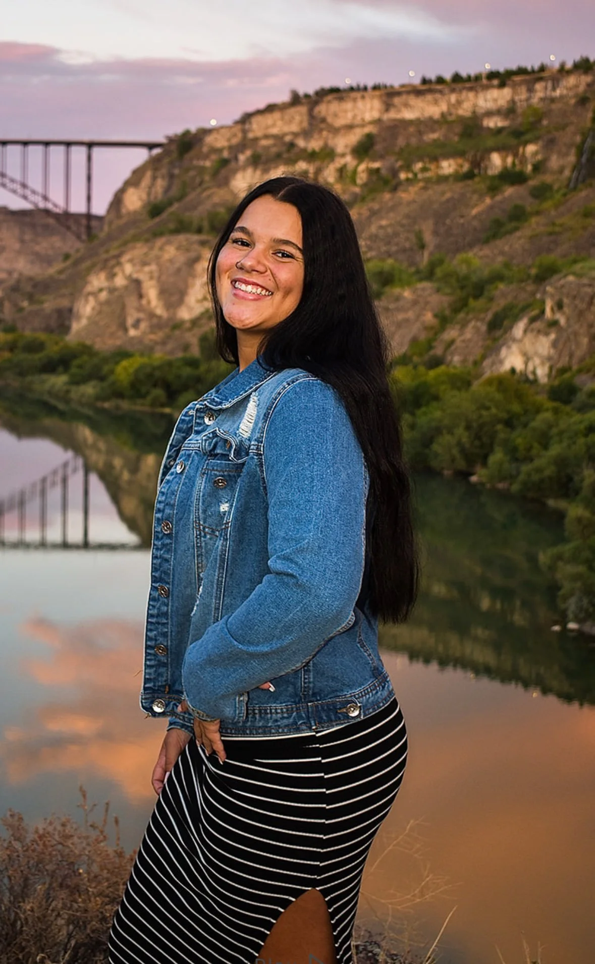 A young woman smiling and posing outdoors at sunset near a body of water with mountainous landscape and a bridge in the background, wearing a denim jacket and striped skirt.