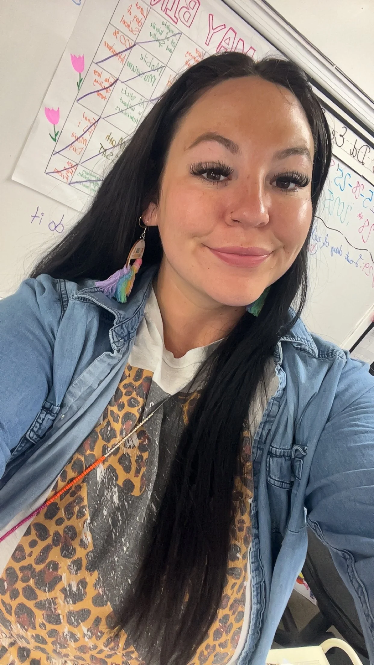 A woman with long black hair, wearing colorful earrings and a denim jacket, smiling in front of a whiteboard with writing and drawings.