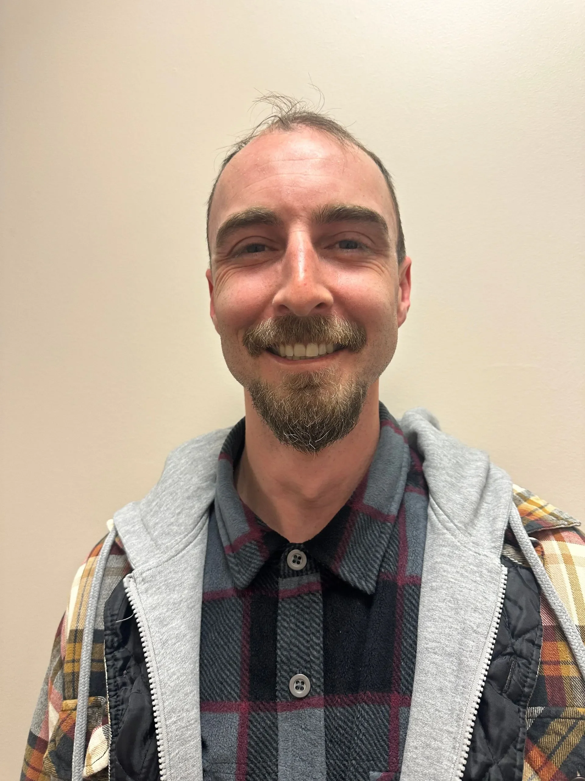 A smiling man with a beard and mustache, wearing a plaid shirt, a gray hoodie, and a jacket, standing against a beige wall.