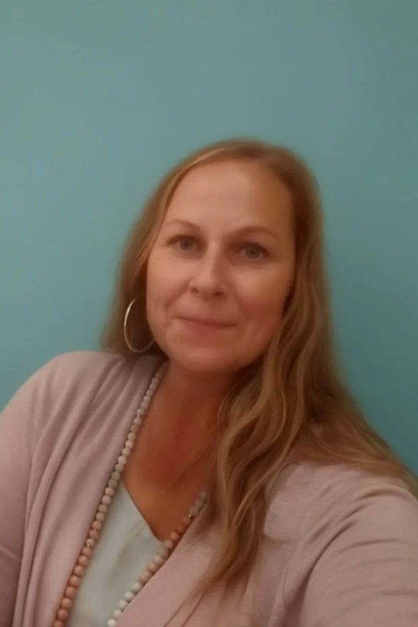 A woman with long light brown hair and hoop earrings, wearing a light-colored cardigan over a white top with a beaded necklace, standing against a blue background.
