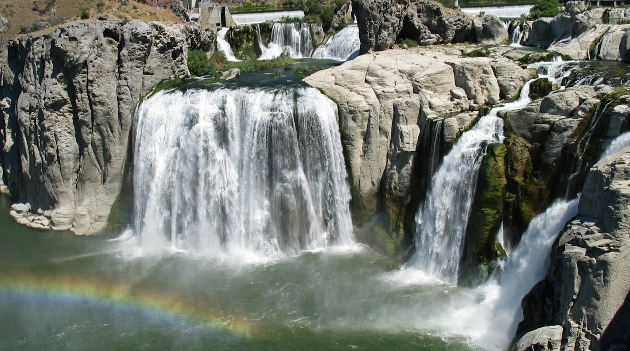 Multiple waterfalls cascading over rocks into a river, with a rainbow visible in the mist at the bottom of the waterfalls.