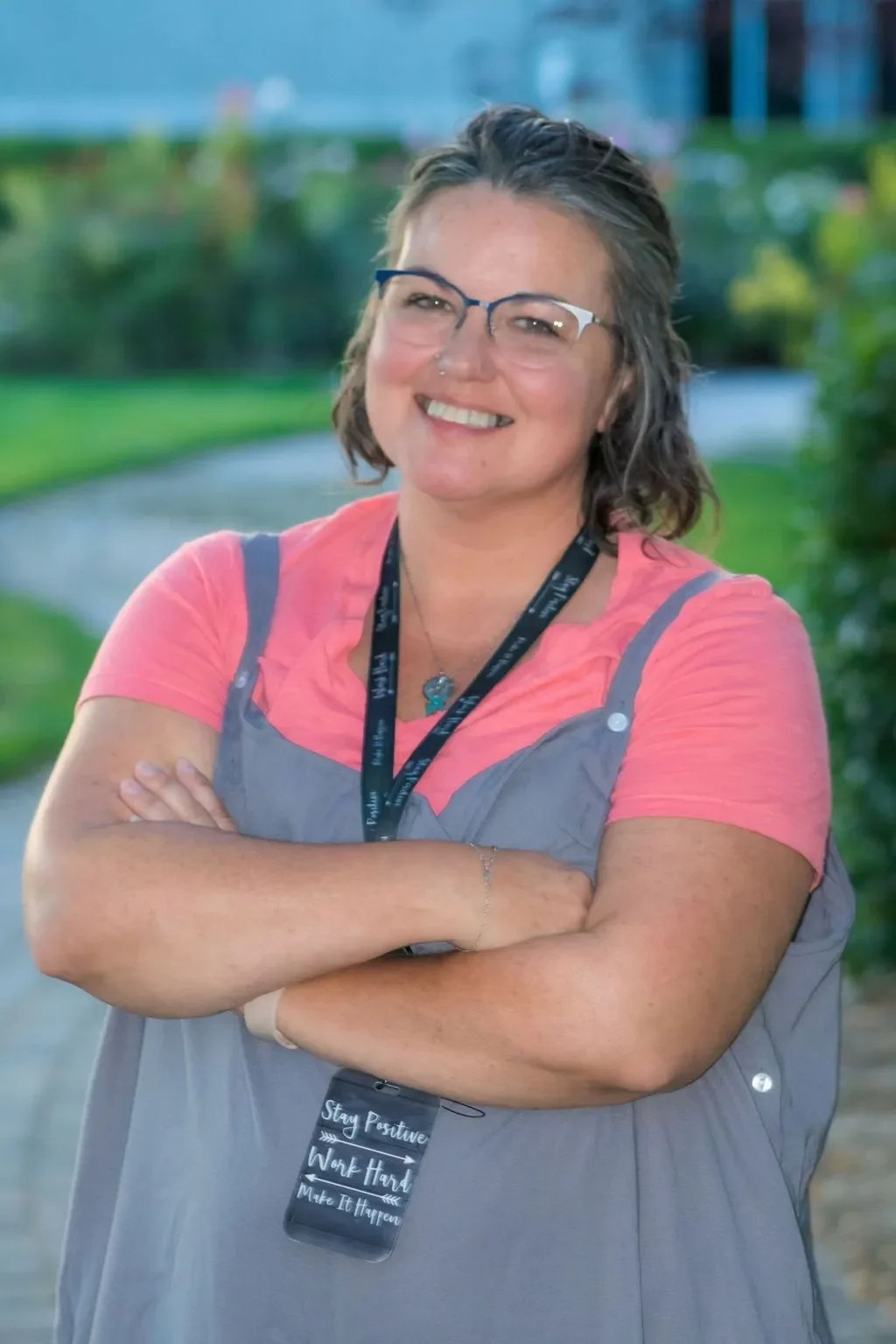 A smiling woman with glasses and curly short hair, wearing a pink t-shirt and gray overalls, standing outdoors on a walking path with greenery in the background.