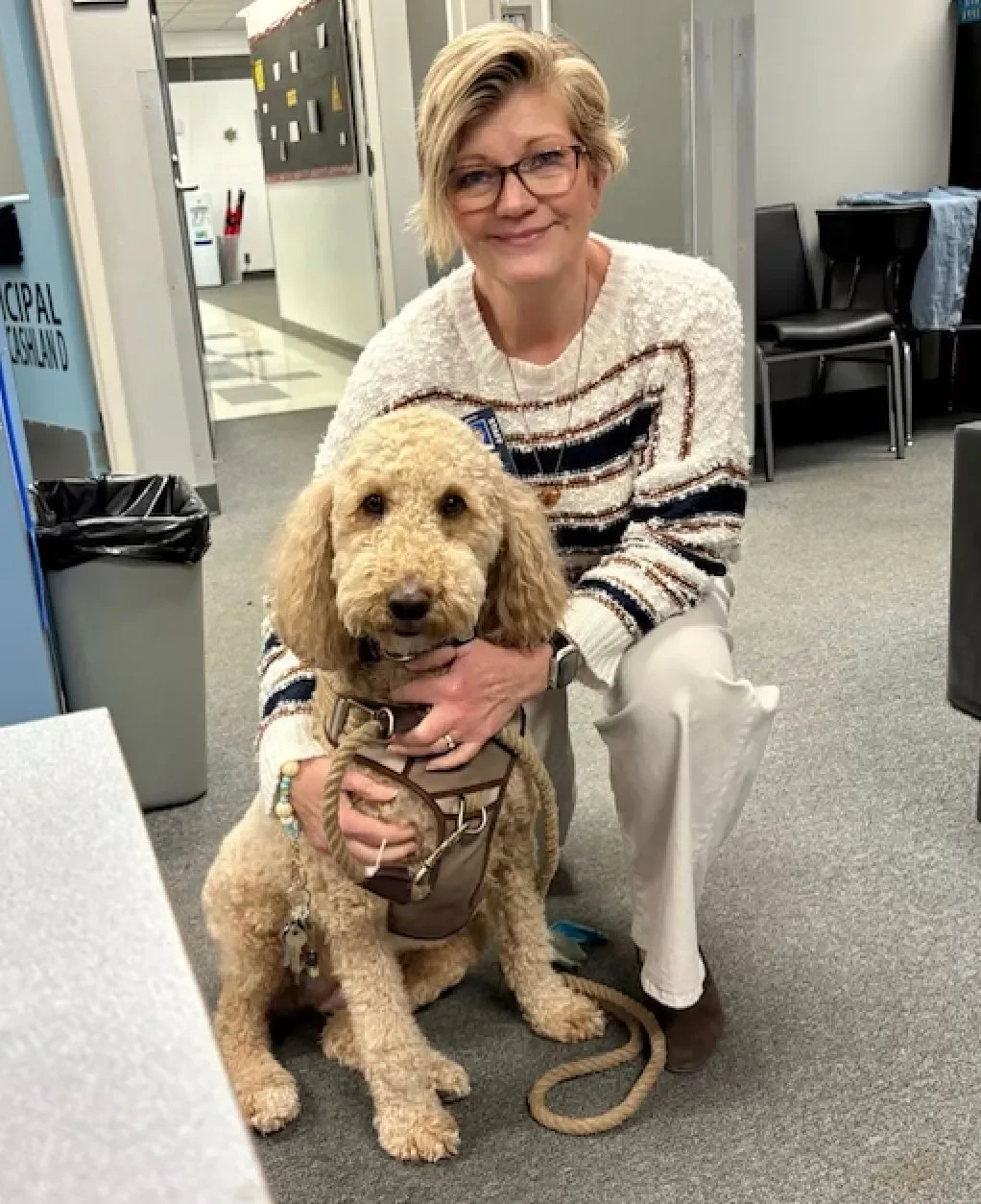 A woman with short blonde hair, glasses, and a striped sweater kneeling next to a service dog in an office setting.