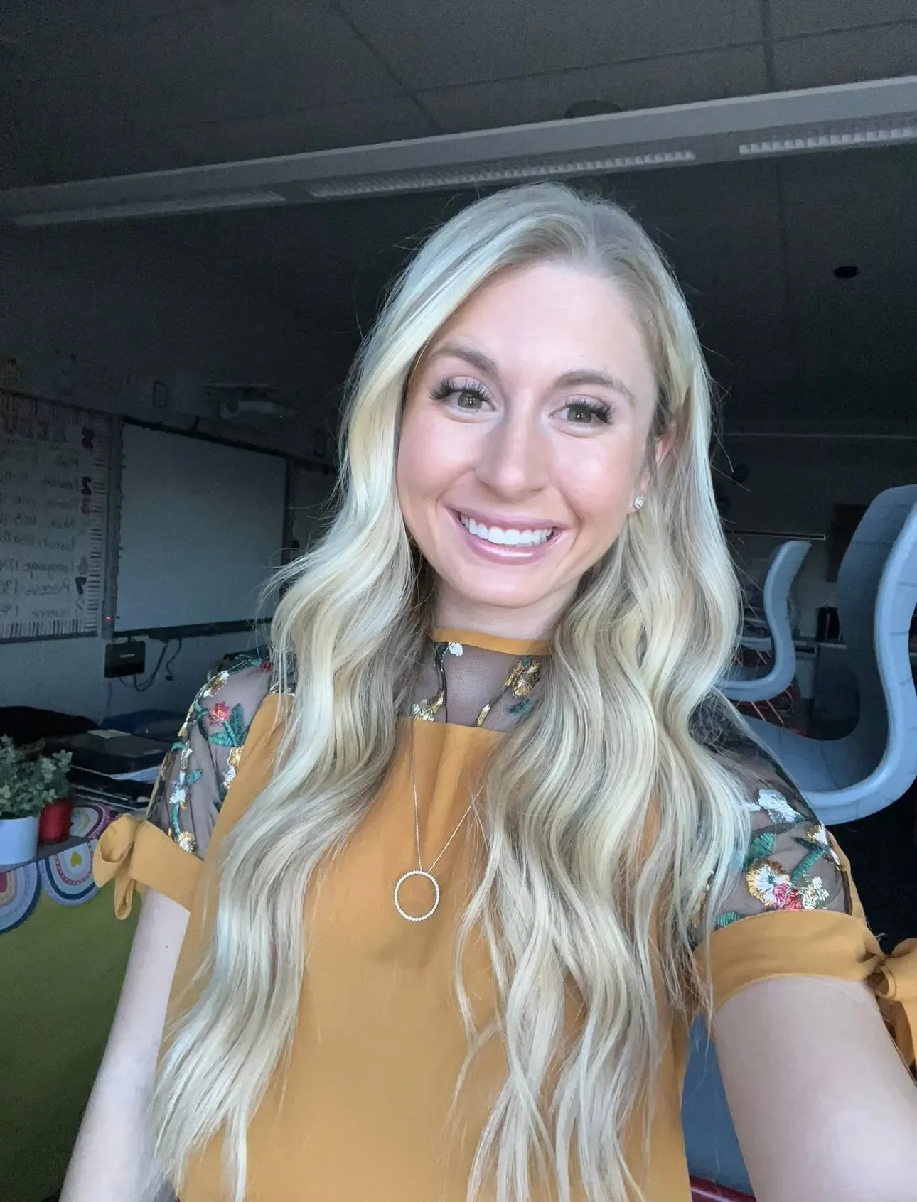 Young woman with long blonde wavy hair taking a selfie in an office or classroom setting, smiling at the camera.