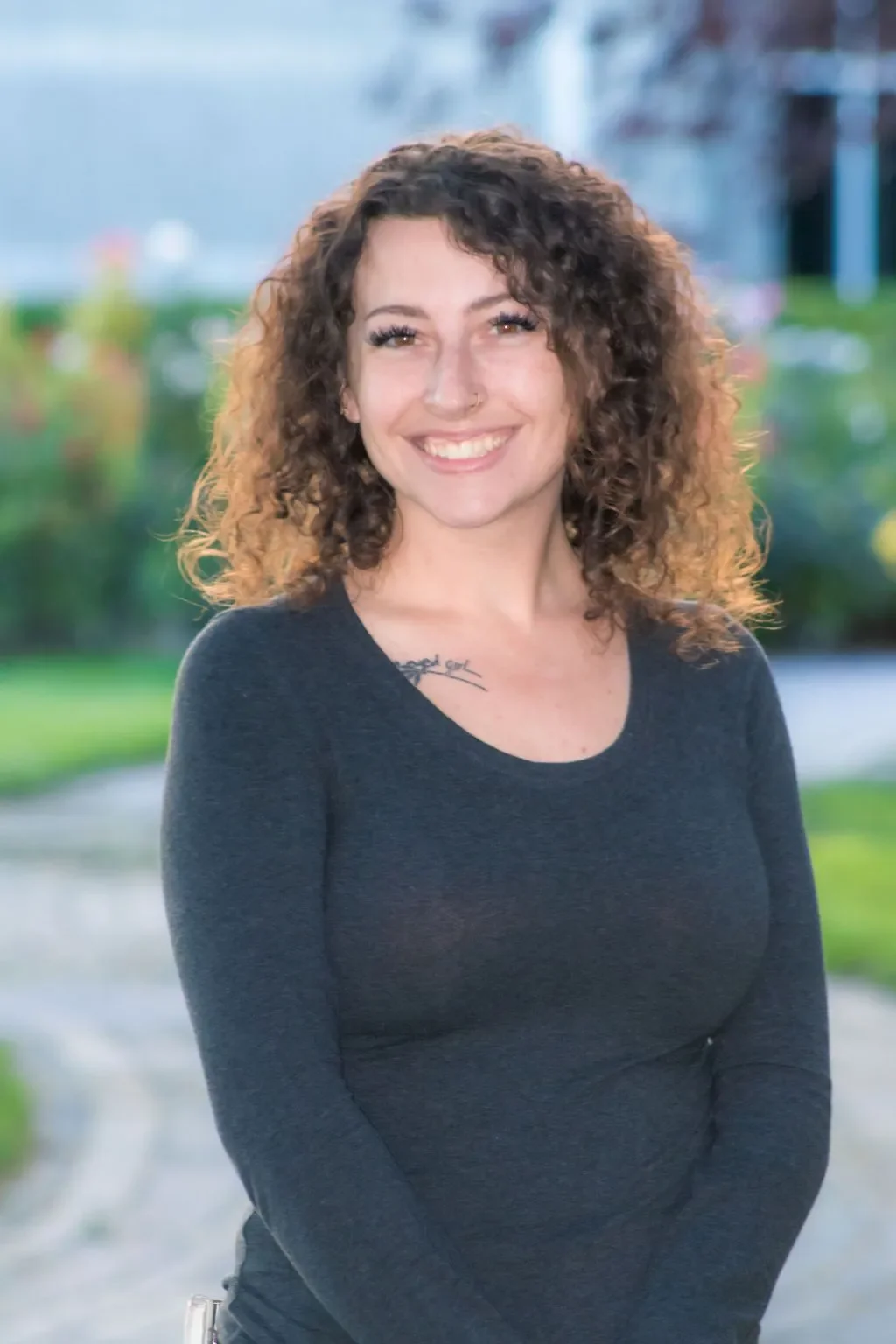 A smiling woman with curly brown hair, wearing a dark long-sleeve top, standing outdoors with a blurred background of greenery and a building.