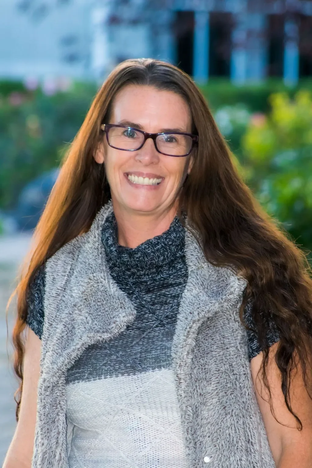A woman with long brown hair, glasses, and a multicolored gray, black, and white vest smiling outdoors with greenery and blurred buildings in the background.