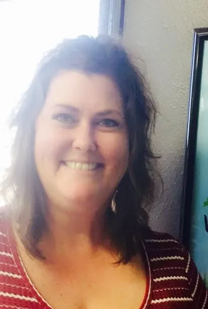 A woman with wavy brown hair smiling indoors near a computer monitor.