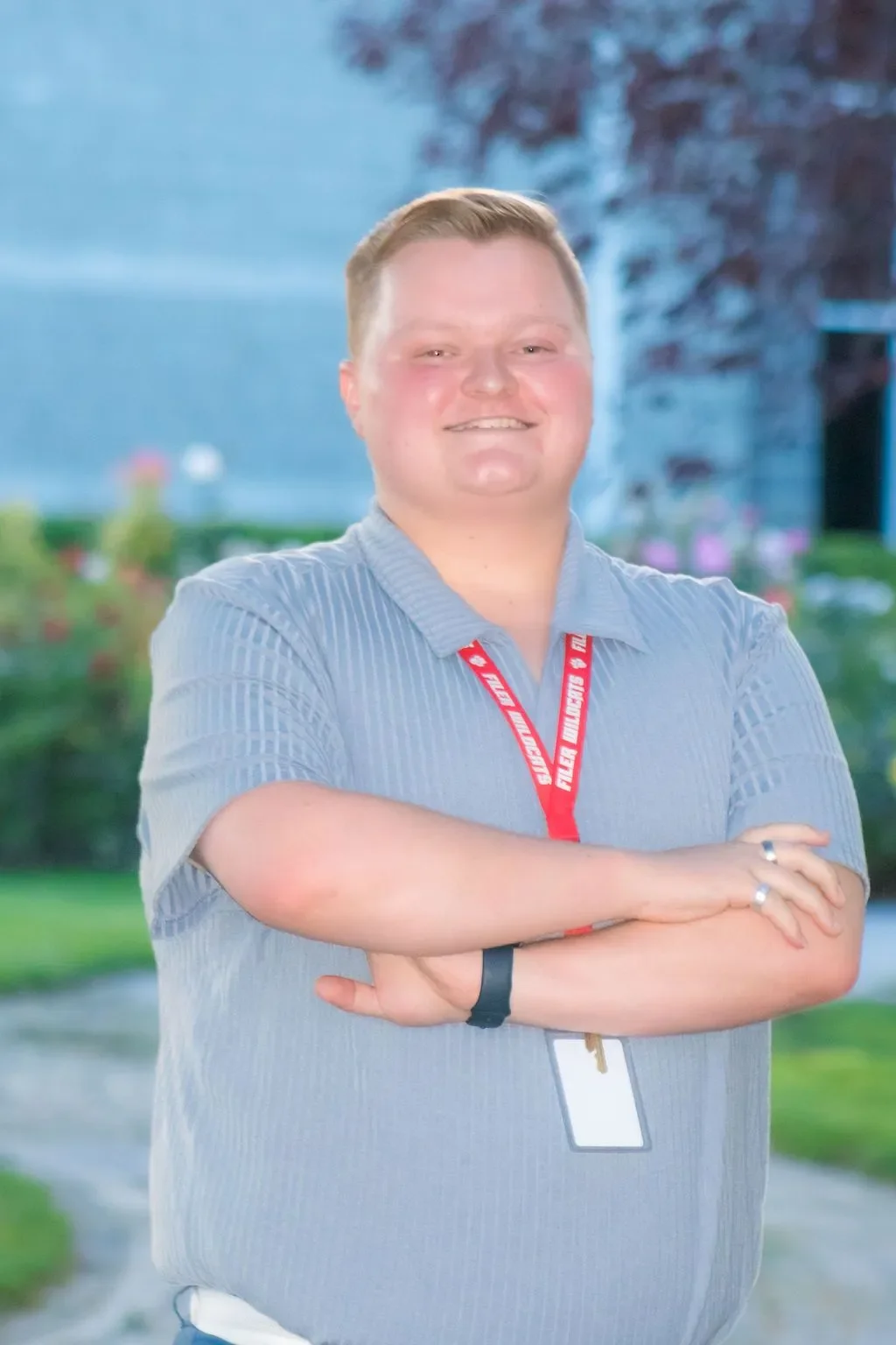 A man smiling with arms crossed, standing outdoors with greenery and trees in the background, wearing a gray short-sleeved shirt, a red lanyard, and a watch.
