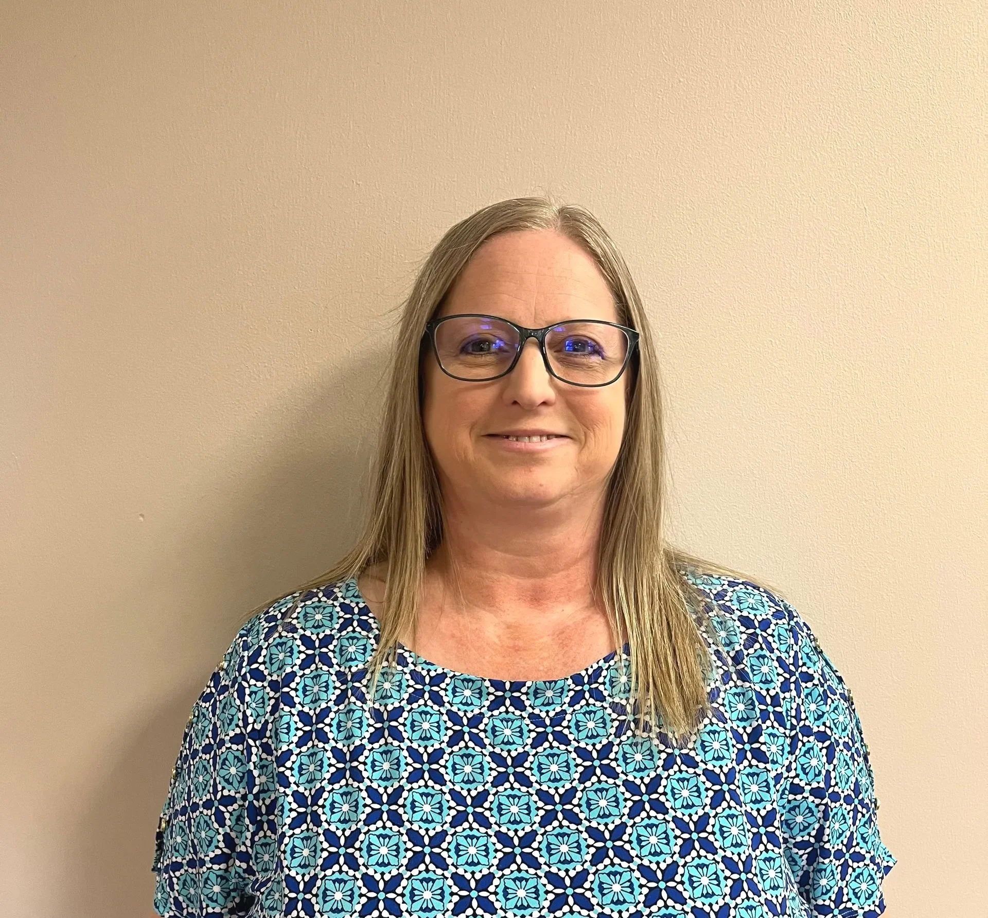 A middle-aged woman with shoulder-length blonde hair, wearing glasses and a blue patterned blouse, smiling while standing against a plain beige wall.