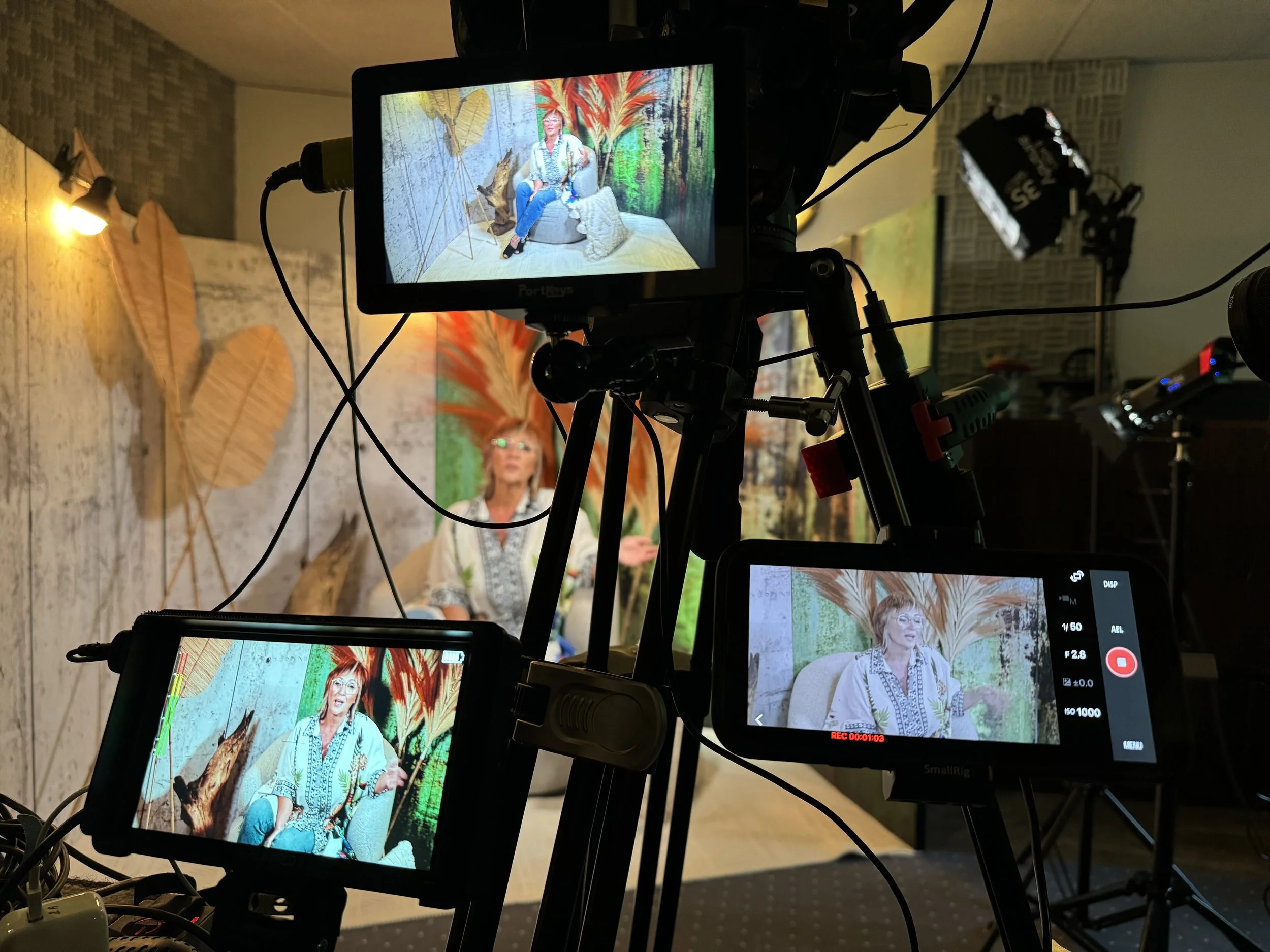Een opnamestudio met drie camera's gericht op een vrouw die in een kleurrijk decor spreekt. De achtergrond heeft grote bladeren en felgekleurde decorstukken. De vrouw zit op een beige stoel en draagt een krullend kapsel, een blouse en een ketting.