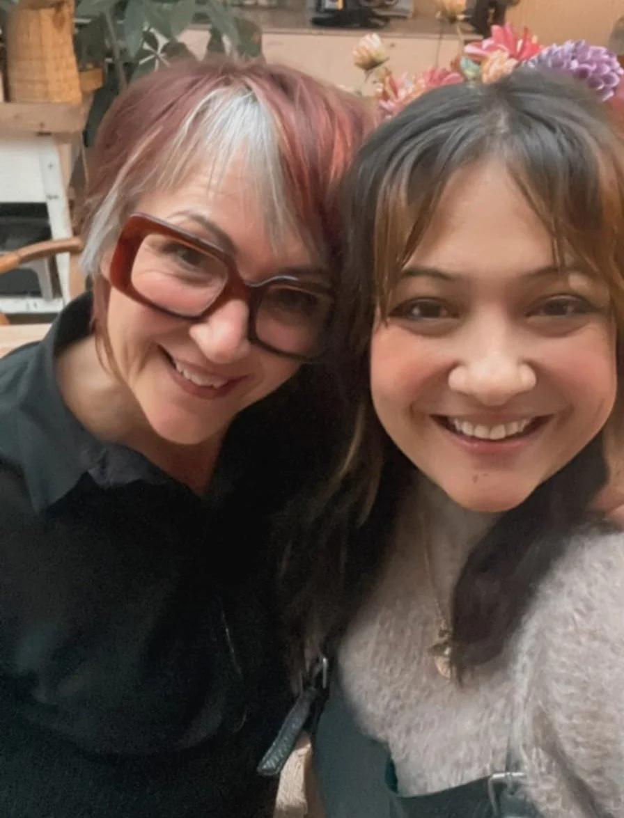 Twee vrouwen lachen en poseren samen voor de camera, de ene draagt grote rode brillen en de andere heeft een bloemenkrans op haar hoofd.