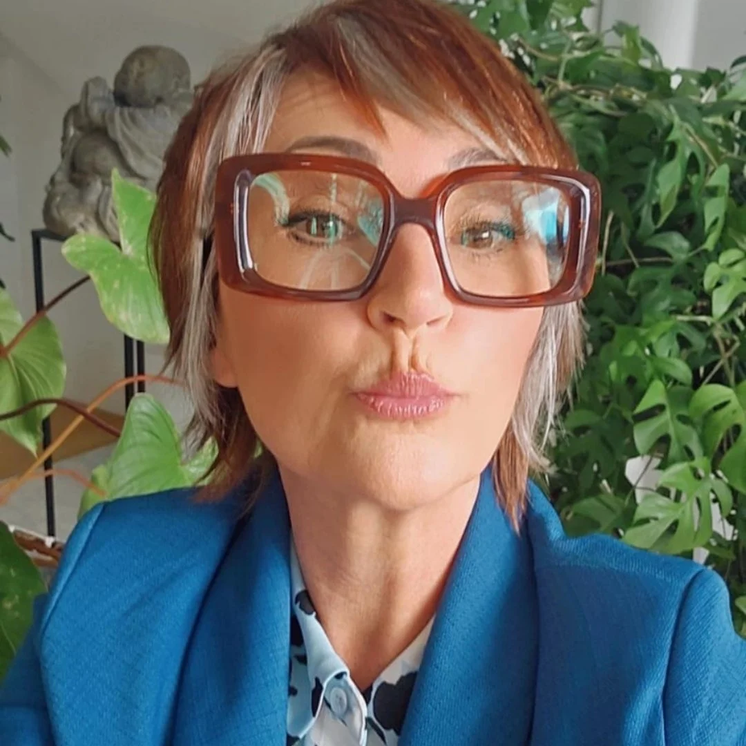 Een vrouw met kort bruin haar en grote bruine brillen die in een groene omgeving staat, met planten op de achtergrond.